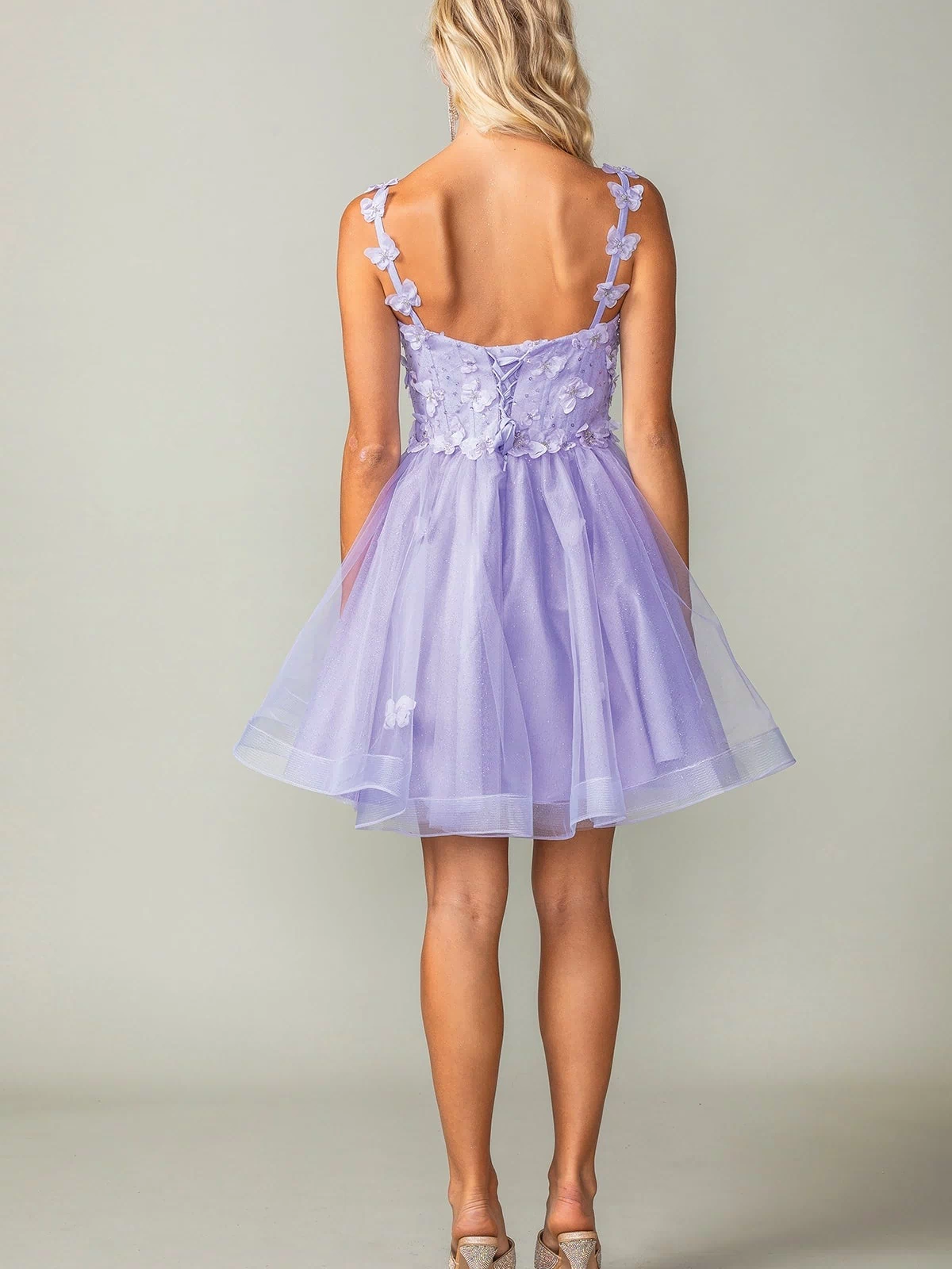 A woman faces away, wearing the Lace-Up Short Butterfly Homecoming Dress 3369 in lavender with floral accents, 3-D butterflies, sheer layers, and high heels.