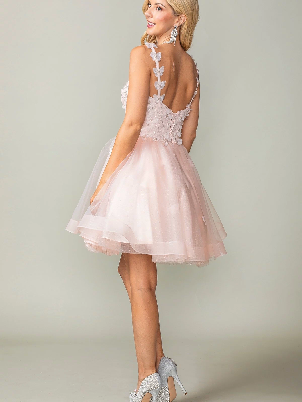 Woman in a Lace-Up Short Butterfly Homecoming Dress 3369, featuring pale pink layered tulle, 3-D butterflies, and a sleeveless design, wears silver heels and stands looking over her shoulder.