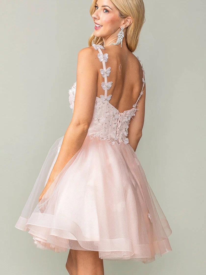 Wearing the Lace-Up Short Butterfly Homecoming Dress 3369, a woman in a light pink sleeveless dress with 3-D butterflies and floral appliqués stands slightly turned, revealing its open back and layered tulle skirt.