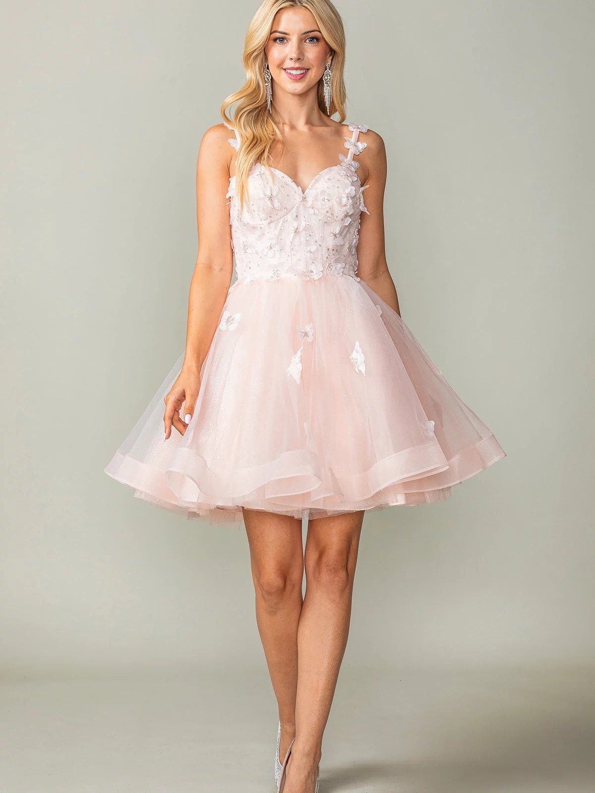 A woman in the Lace-Up Short Butterfly Homecoming Dress 3369 featuring a light pink, sleeveless design with 3-D butterflies and a tulle skirt, paired with silver heels, stands against a plain background.