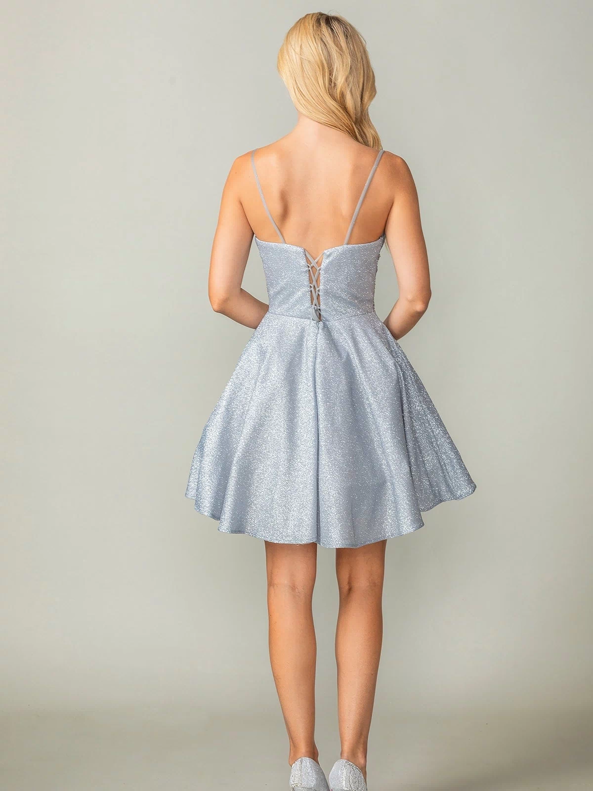 A woman faces away, wearing the Cowl-Neck Short Glitter Homecoming Dress 3386—a sparkly light blue, lace-up mini dress with thin straps—paired with matching high heels.
