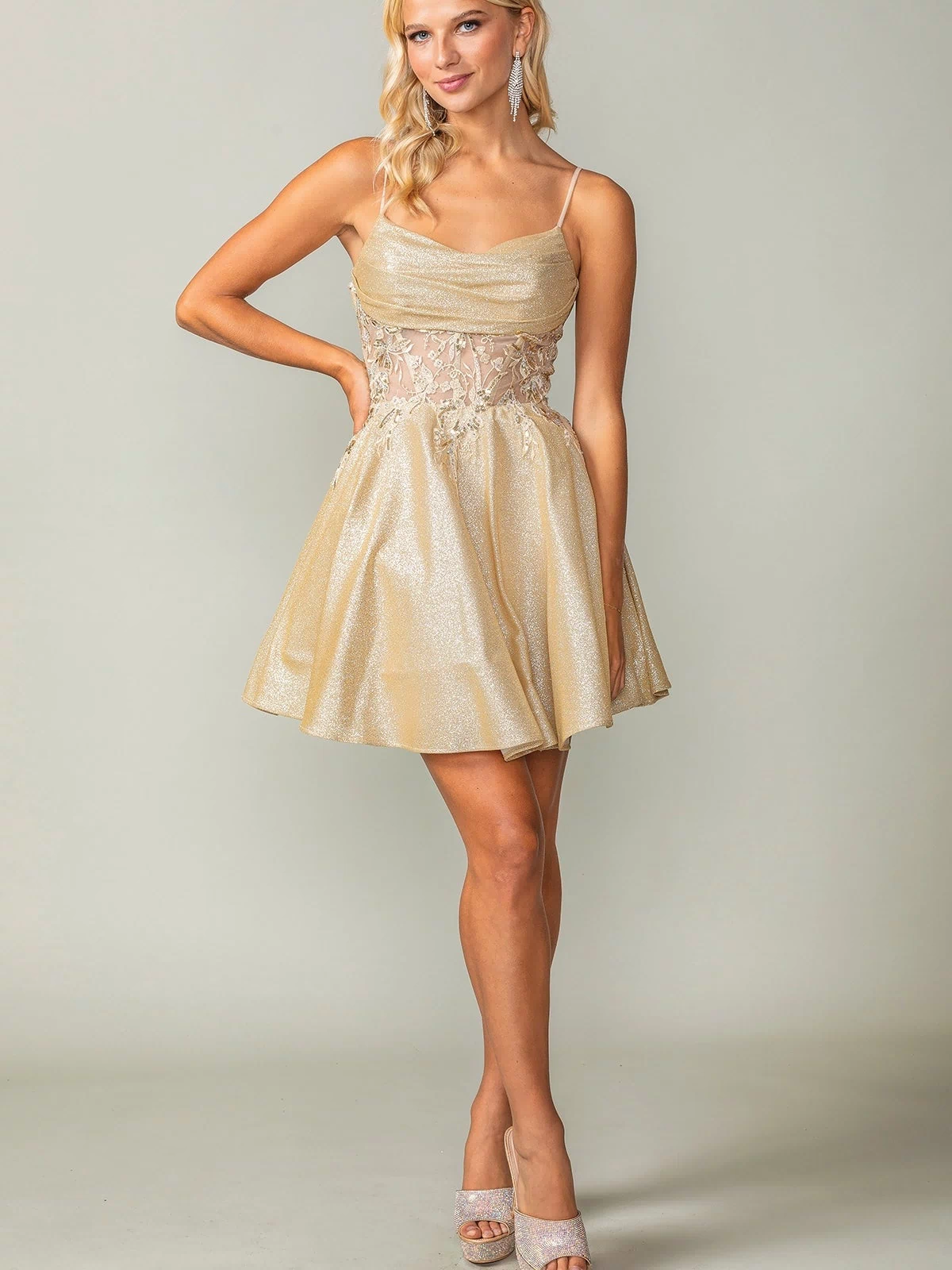 A woman wears the Cowl-Neck Short Glitter Homecoming Dress 3386, featuring sparkly fabric and a stylish silhouette, paired with matching high heels against a plain background.