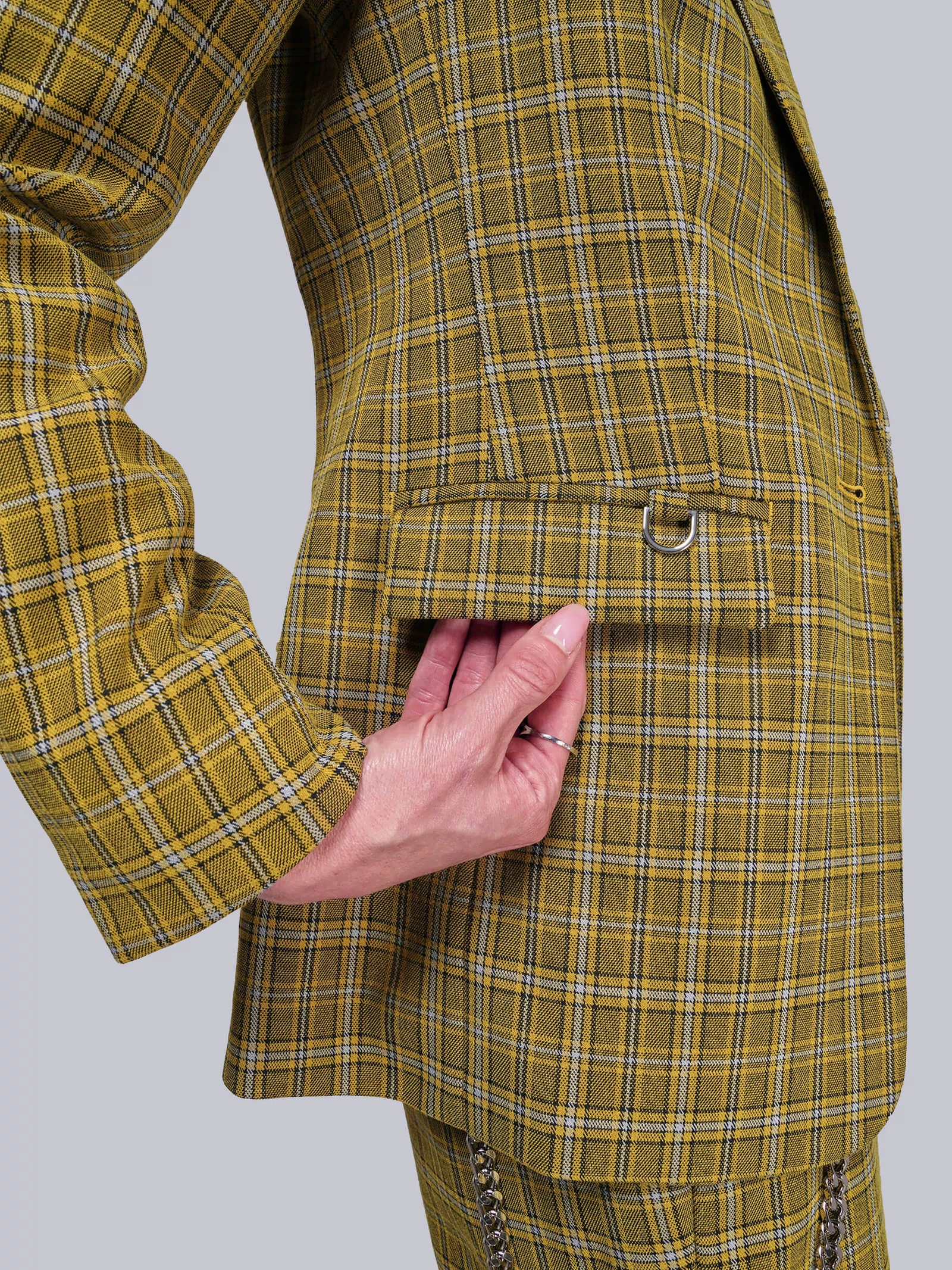 S=Close up of yellow plaid blazer with a hand touching the pocket flap on a light gray background
