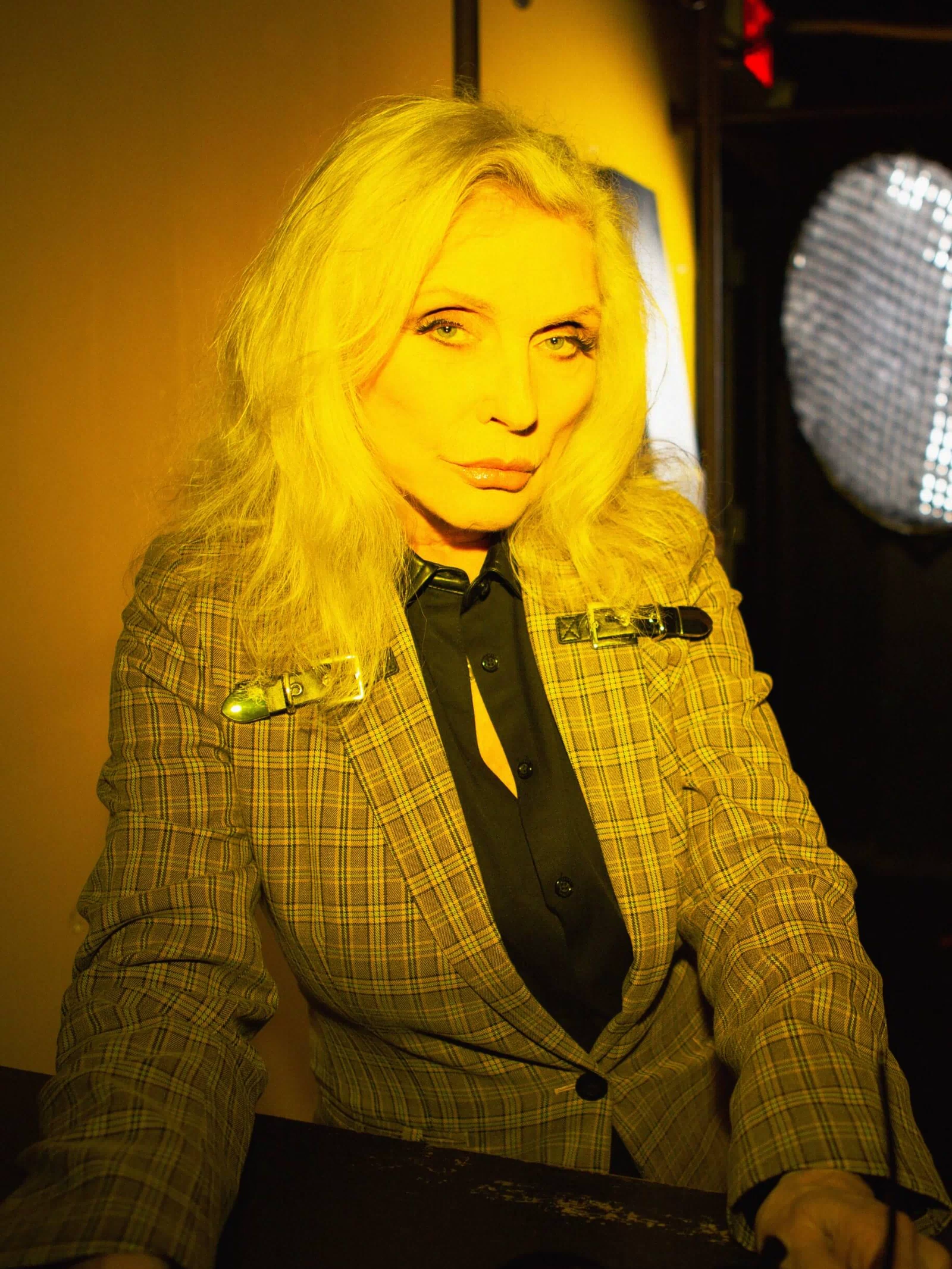 Person with blonde hair wearing a checkered suit in a dimly lit room.