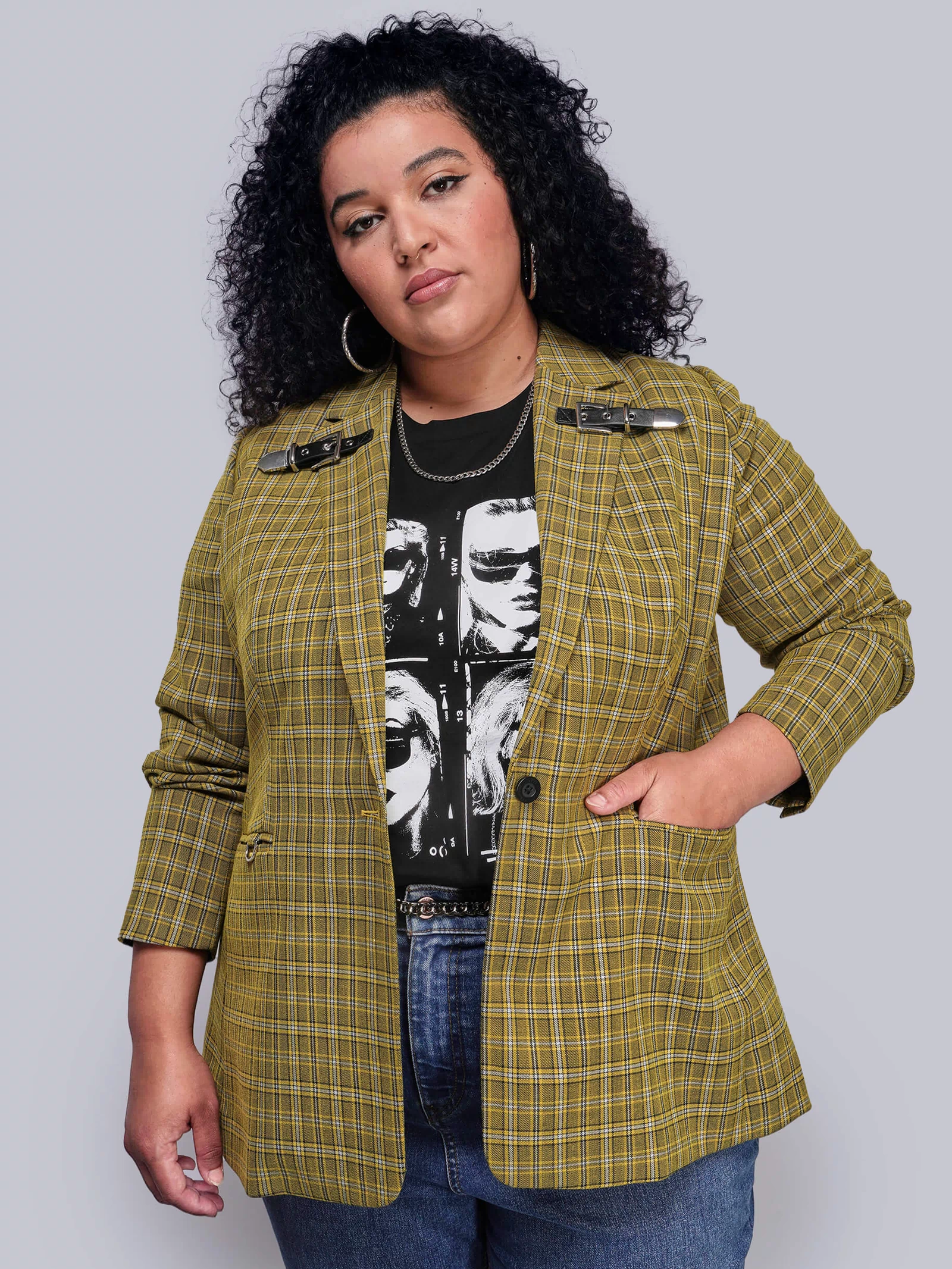 1X=Woman wearing a yellow plaid blazer over a black graphic t-shirt on a plain background