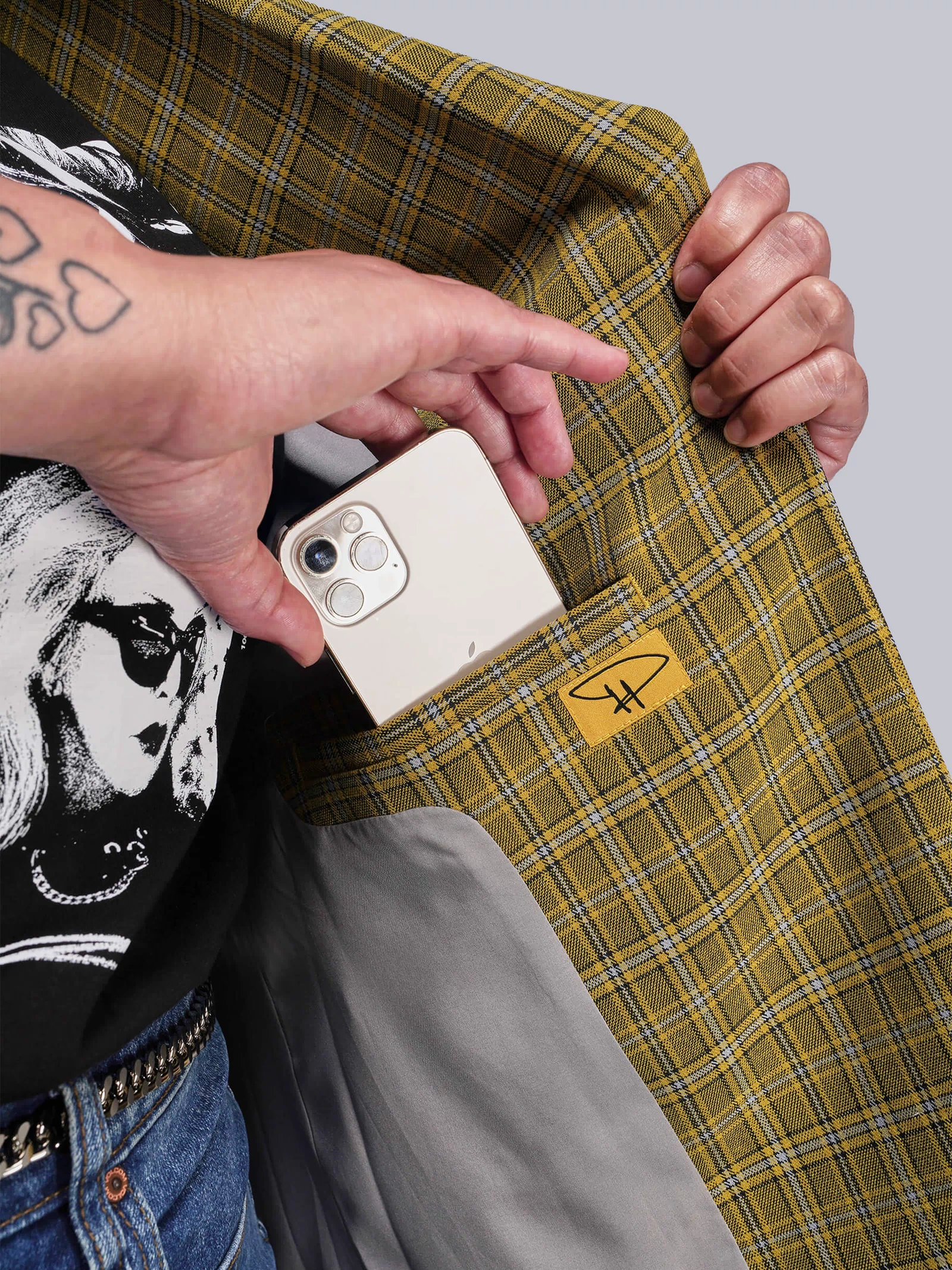 1X=Person holding a phone placing inside the inner pocket of a yellow plaid jacket with a Debbie Harry signature patch.