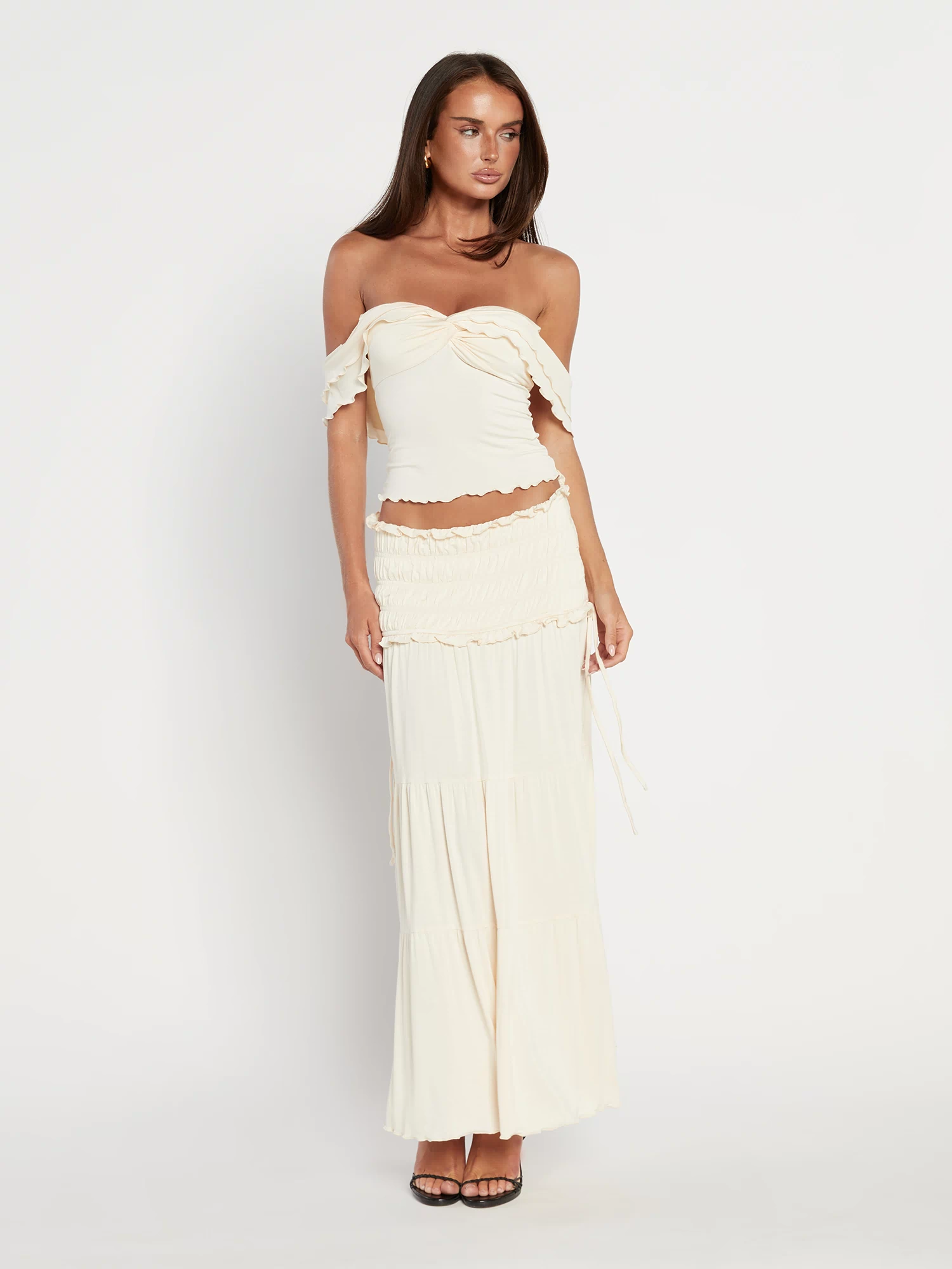 A woman stands in heeled sandals against a white background, wearing the TIGERMIST DENALI MAXI SKIRT in Bone with ruffle details and a flared silhouette, paired with a cream off-the-shoulder fitted top.