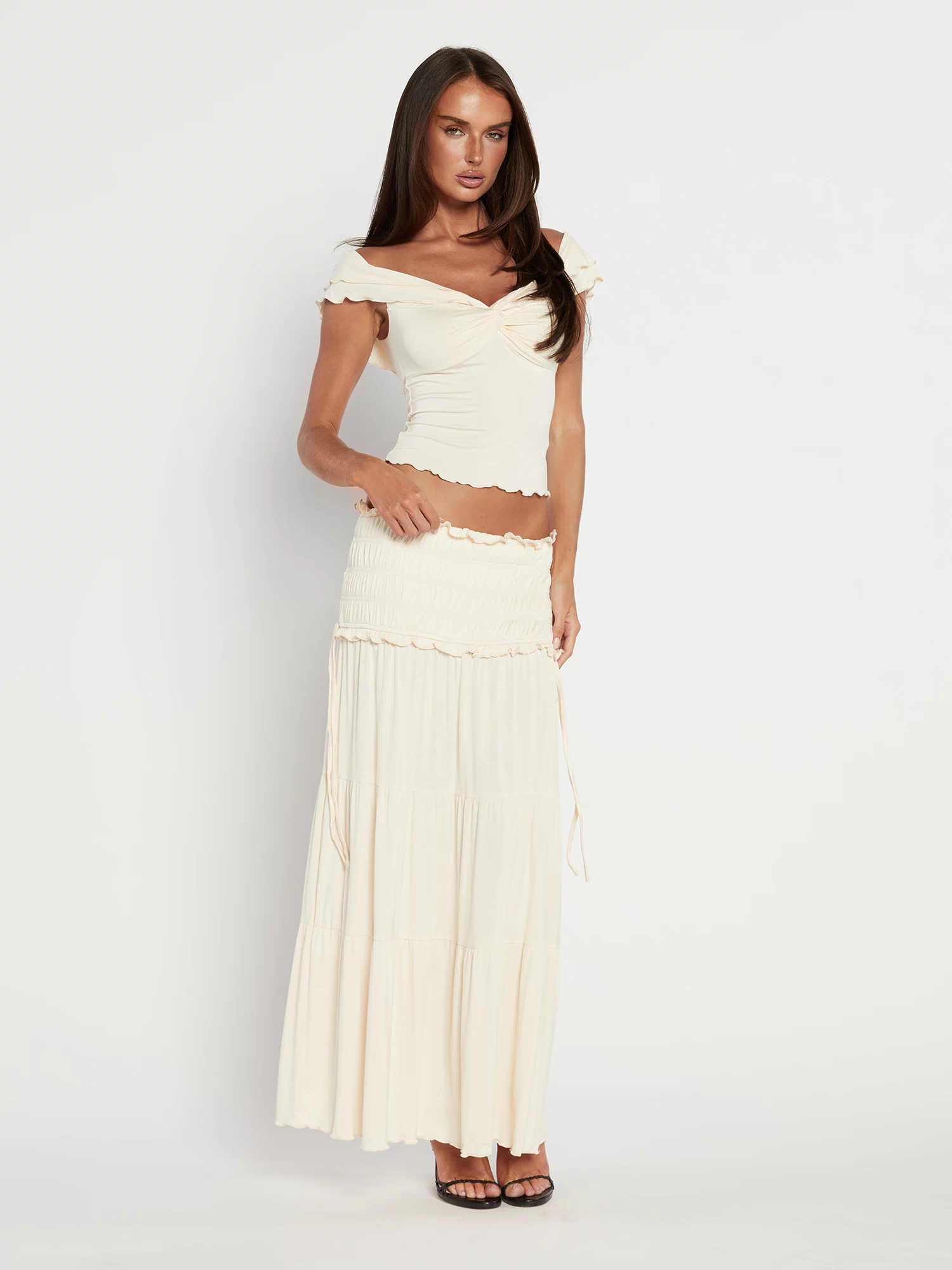 A woman with long brown hair poses against a white background wearing the TIGERMIST DENALI MAXI SKIRT - NEUTRAL : BONE, styled with an off-shoulder cream crop top and black high-heeled sandals. The skirt features a flared silhouette and ruffled edges.