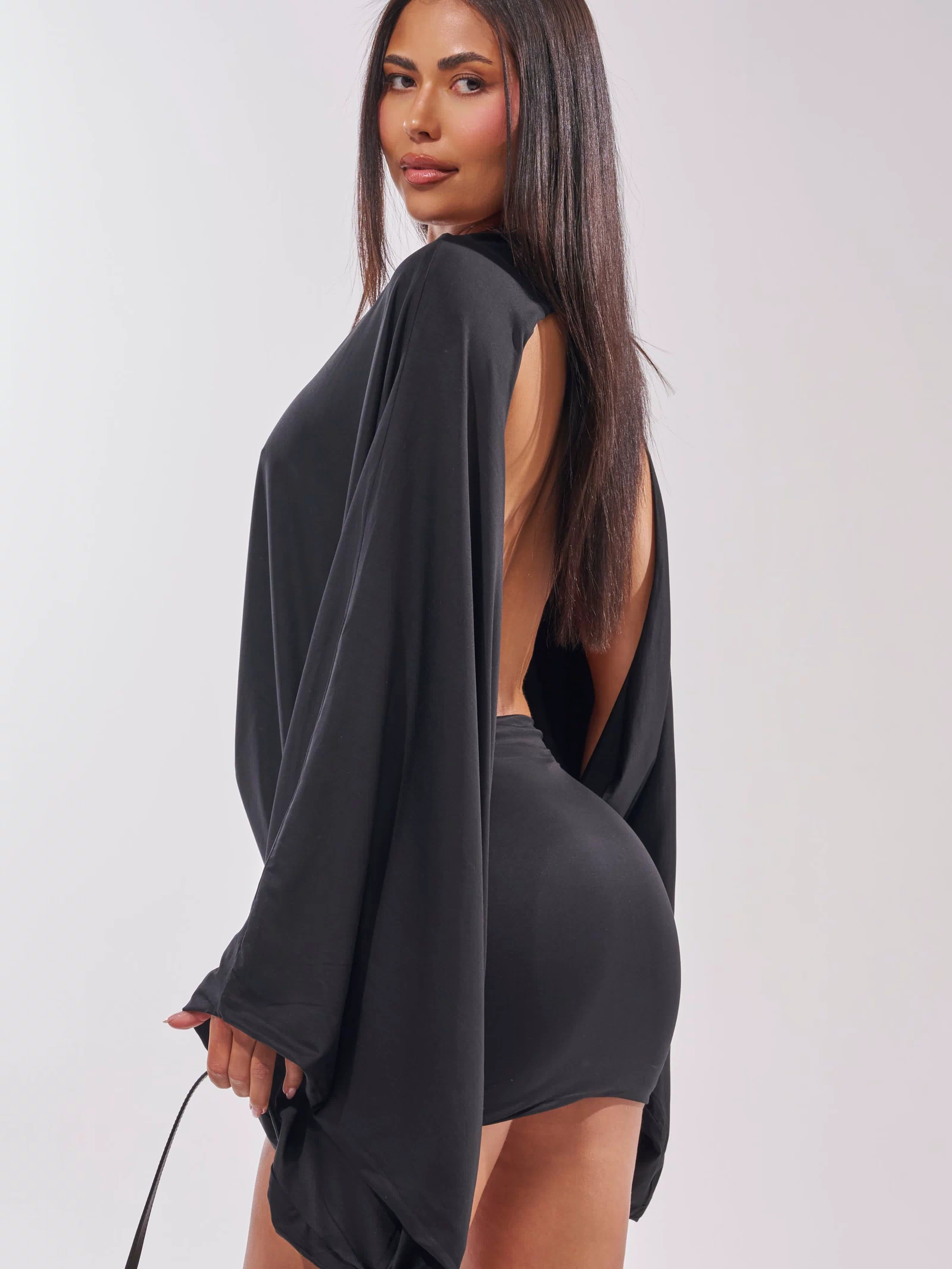A woman with long straight hair models the DIANA DRAPED MINI DRESS IN BLACK, featuring batwing sleeves and an open back, as she poses and looks over her shoulder against a plain light background.