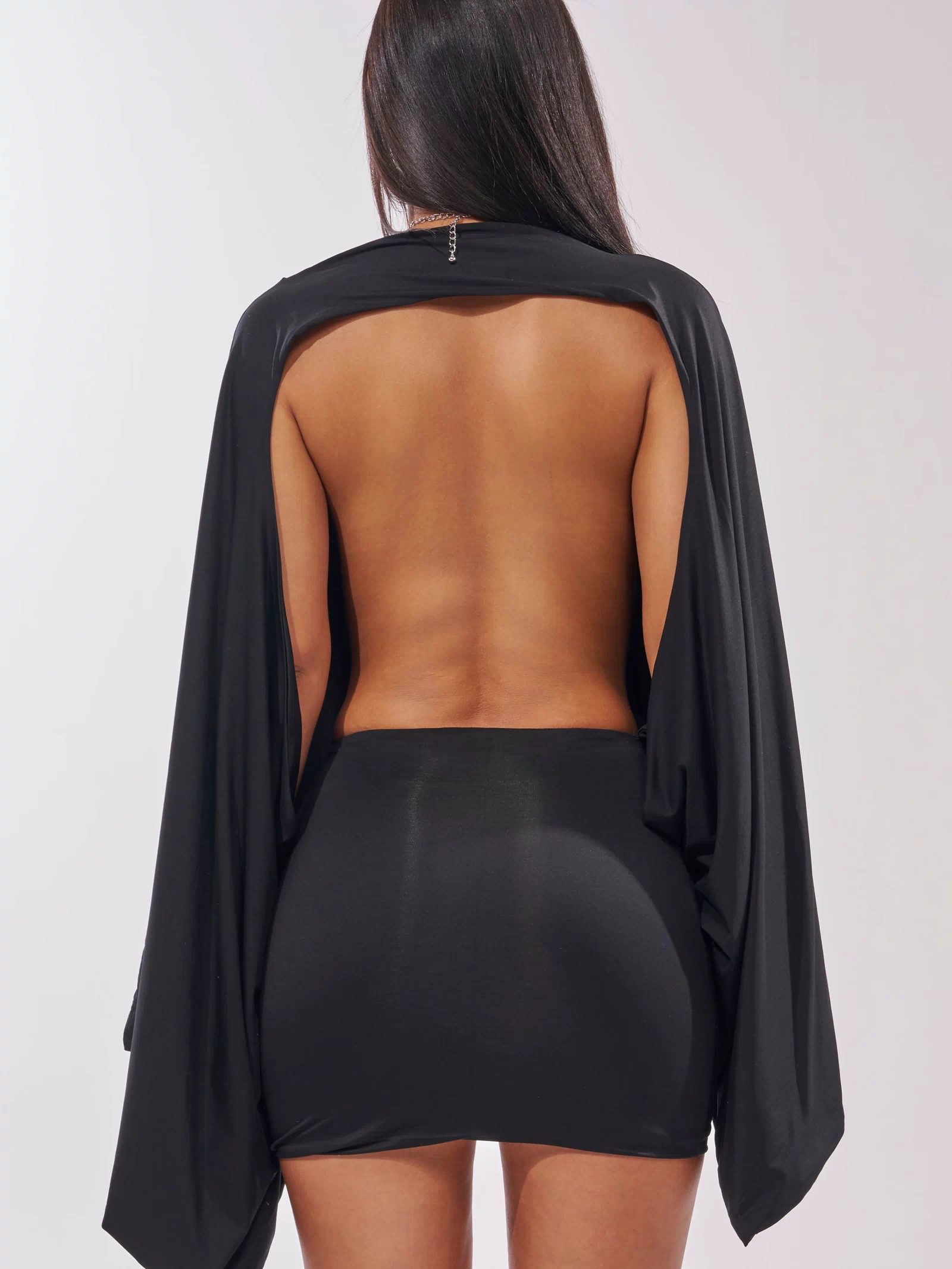 A woman faces away from the camera wearing the DIANA DRAPED MINI DRESS IN BLACK, which features batwing sleeves, a fitted mini skirt, and reveals her bare back and arms.