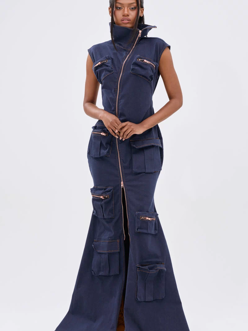 A woman faces forward in the DONT MESS WITH ME COLLARED DENIM MAXI DRESS, a dark wash, sleeveless maxi with a high collar, front zipper from neck to hem, and utility pockets with zippers and flaps for a modern, functional style.
