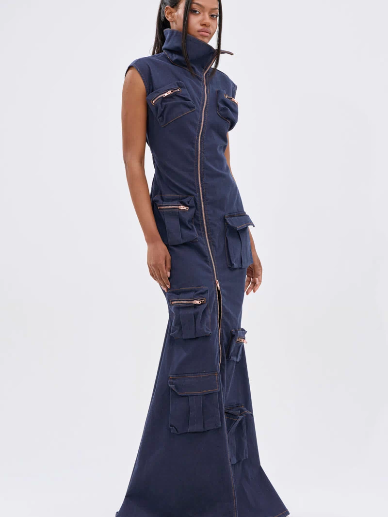 A woman stands against a white background wearing the DONT MESS WITH ME COLLARED DENIM MAXI DRESS, a sleeveless dark wash denim maxi with a high collar, front zipper, and multiple utility pockets on the bodice and skirt.