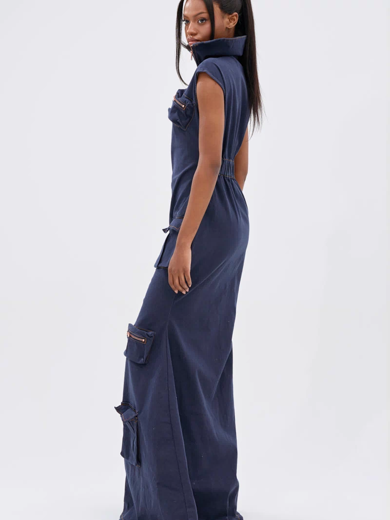 A woman with long straight hair in a high ponytail wears the DONT MESS WITH ME COLLARED DENIM MAXI DRESS, a sleeveless navy blue piece with utility pockets, standing sideways and looking at the camera.