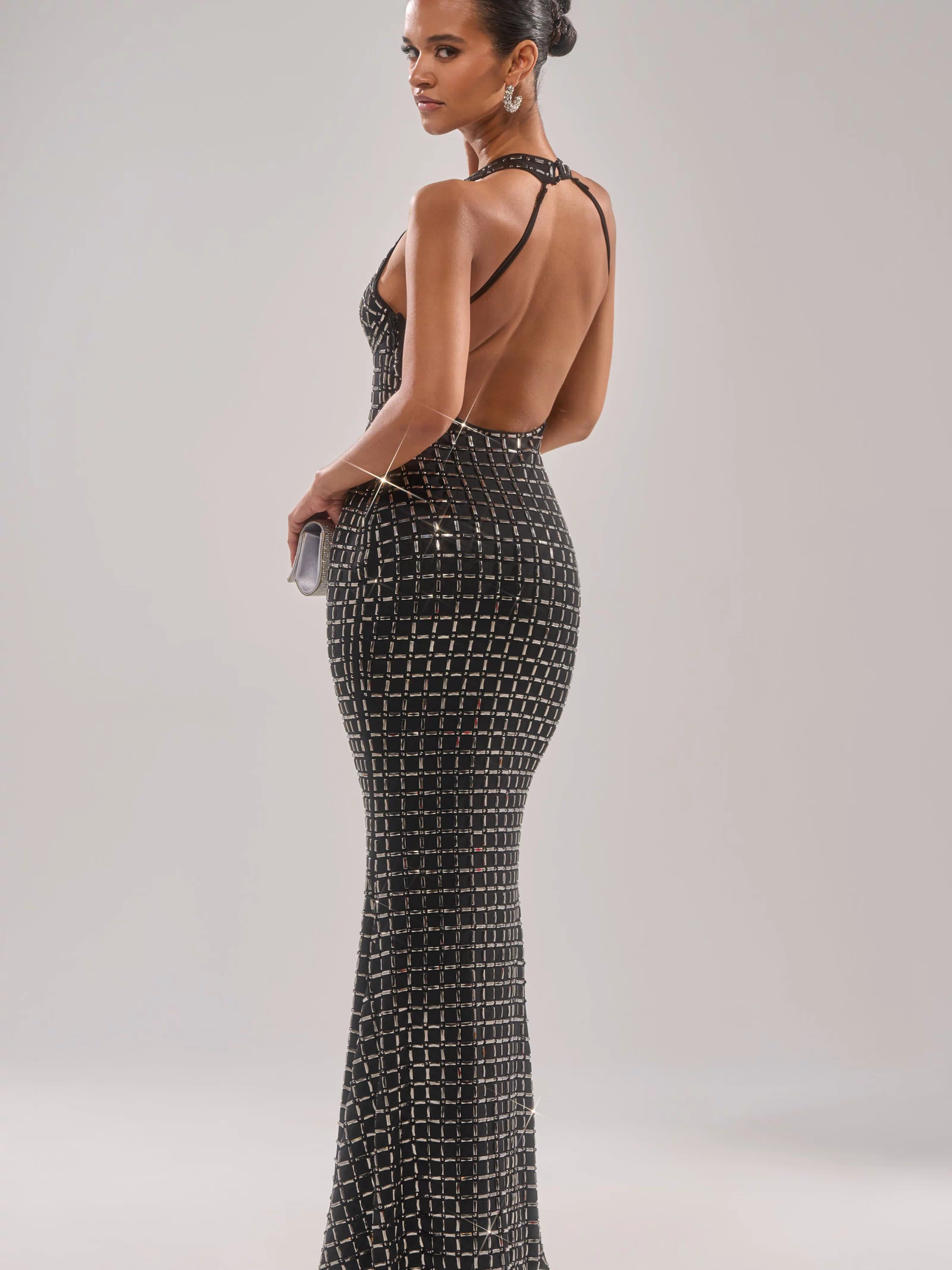 A woman in the DRESSED UP WITH NOWHERE TO GO RHINESTONE MAXI DRESS—floor-length, black with silver geometric rhinestones and a halter neckline—stands hand on hip, looking over her shoulder. She carries a clutch and wears her hair in a sleek bun.
