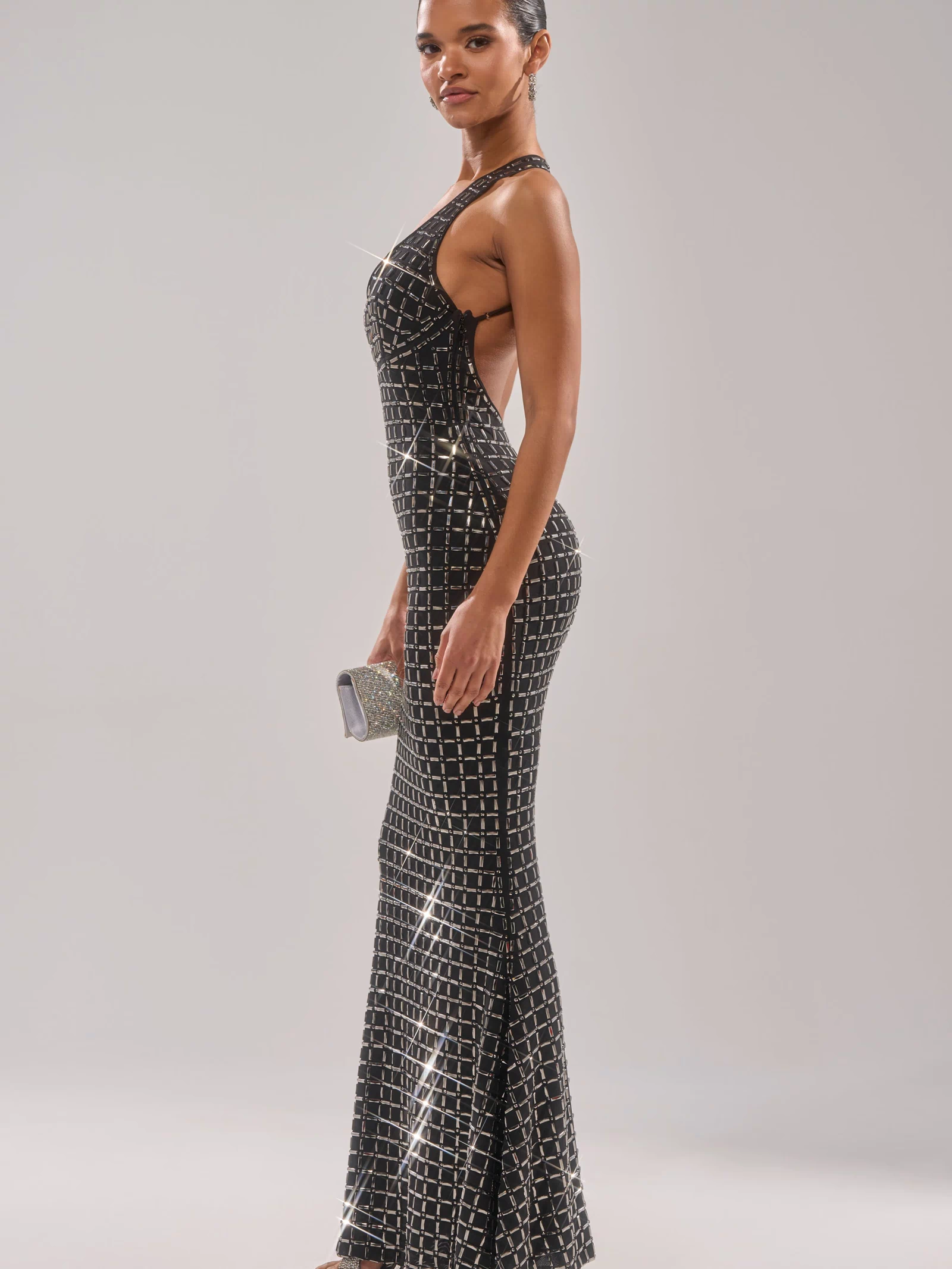 A woman wears the DRESSED UP WITH NOWHERE TO GO RHINESTONE MAXI DRESS, a black formal gown with geometric silver pattern and halter neckline. She holds a small silver clutch and poses in pointed heels against a light background.
