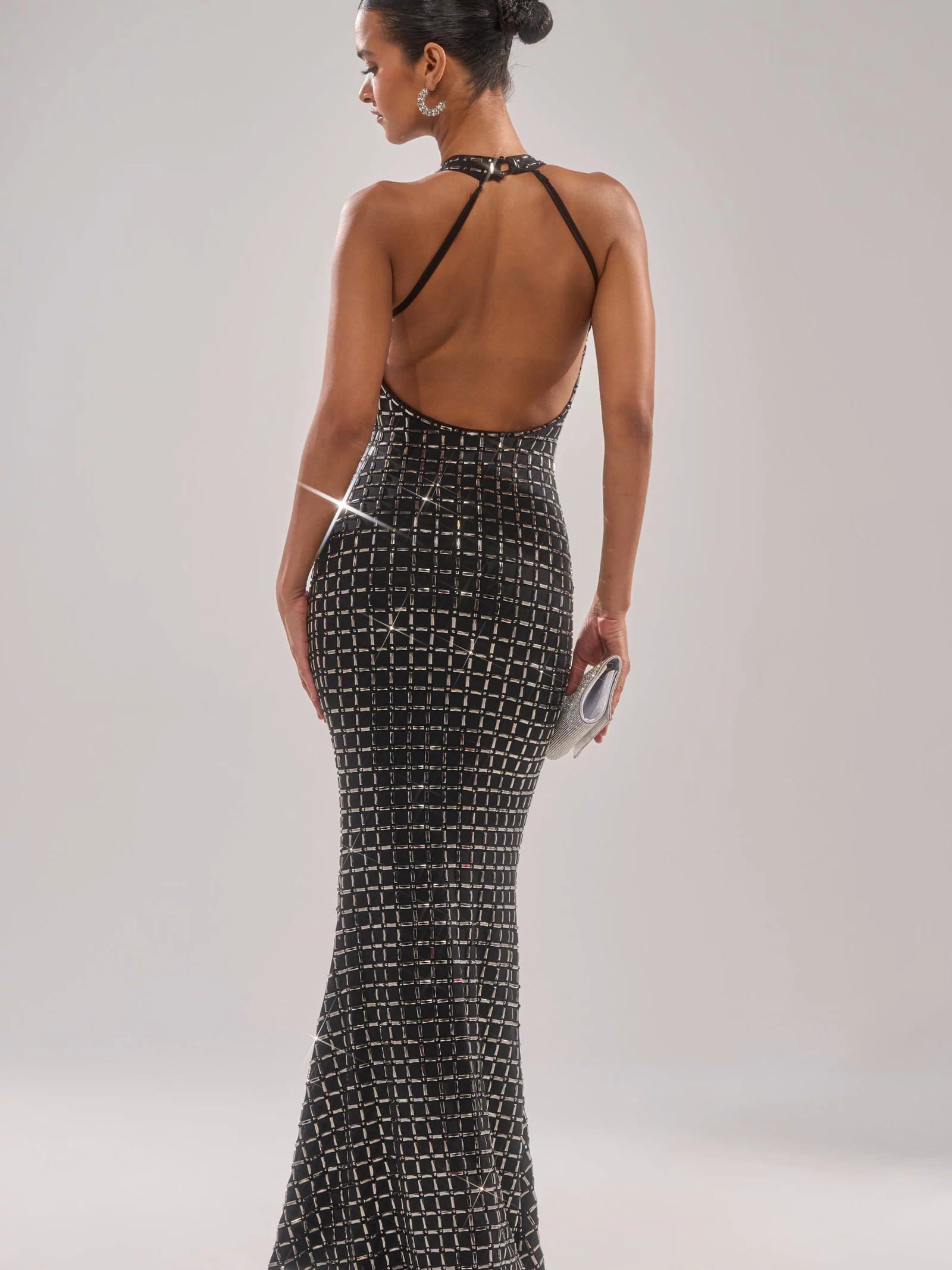 A woman stands facing away in the DRESSED UP WITH NOWHERE TO GO RHINESTONE MAXI DRESS, featuring a sparkling grid pattern, open back, and halter neckline. She holds a clutch and wears her hair in a neat bun.