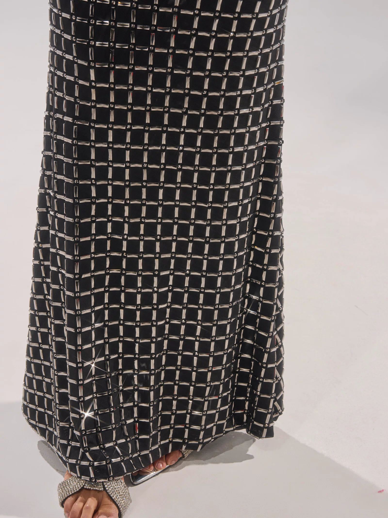 A person stands on a light-colored floor, wearing the DRESSED UP WITH NOWHERE TO GO RHINESTONE MAXI DRESS featuring a black skirt with a white grid pattern and pairing it with open-toe rhinestone sandals. Only lower legs and feet are visible.