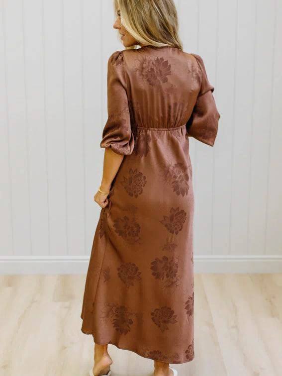 A woman wearing a brown midi dress with a tonal floral jacquard pattern, shown from the back in a bright room with white paneled walls and light wood floors.