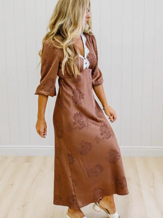 A woman wearing a brown midi dress with a tonal floral pattern, featuring a white lace-up V-neckline detail, standing in a room with light wood floors and white paneled walls.