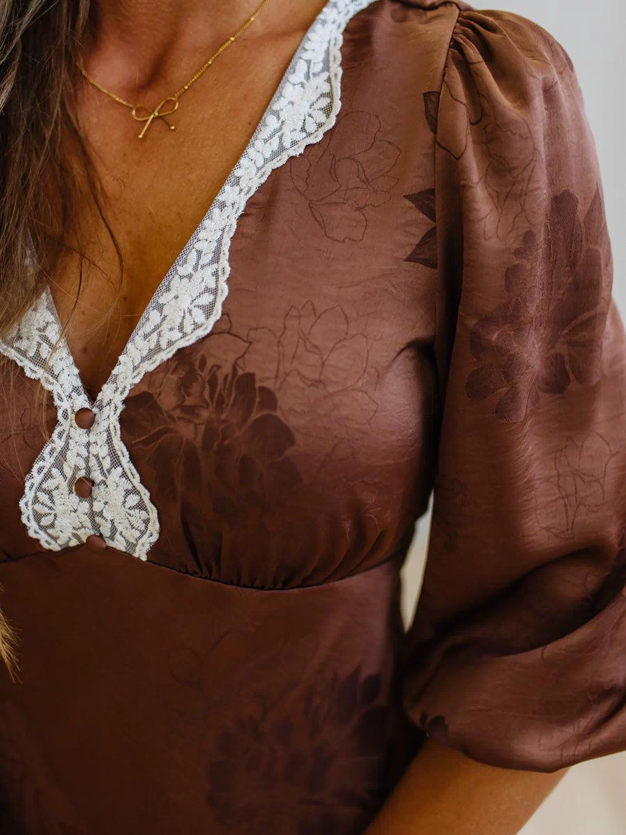 A close-up of a woman wearing a brown satin-like blouse with a floral jacquard pattern, white lace trim along the V-neck and chest, and puff sleeves.