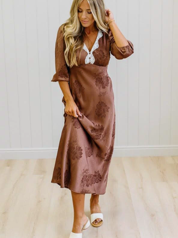 A woman modeling a brown midi-length dress with a subtle floral jacquard pattern, featuring a white lace V-neckline detail, standing on a light wood floor against a white paneled wall.