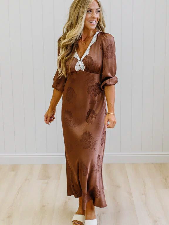A woman modeling a brown satin maxi dress with a tonal floral pattern, featuring a lace V-neckline and puff sleeves, standing in front of a white paneled wall and light wood floor.