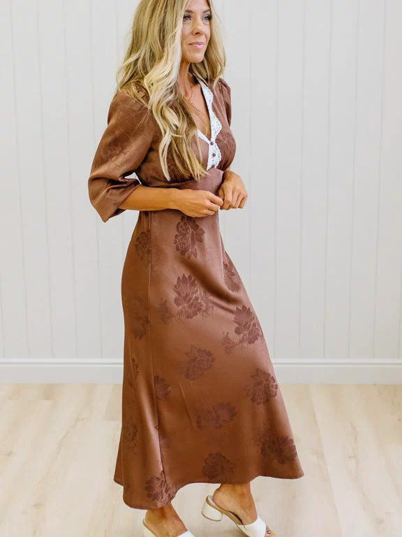 A woman modeling a brown satin-like midi dress with a floral jacquard pattern, featuring a wrap-style collar and three-quarter sleeves, paired with white slide sandals against a white paneled wall and light wood floor.