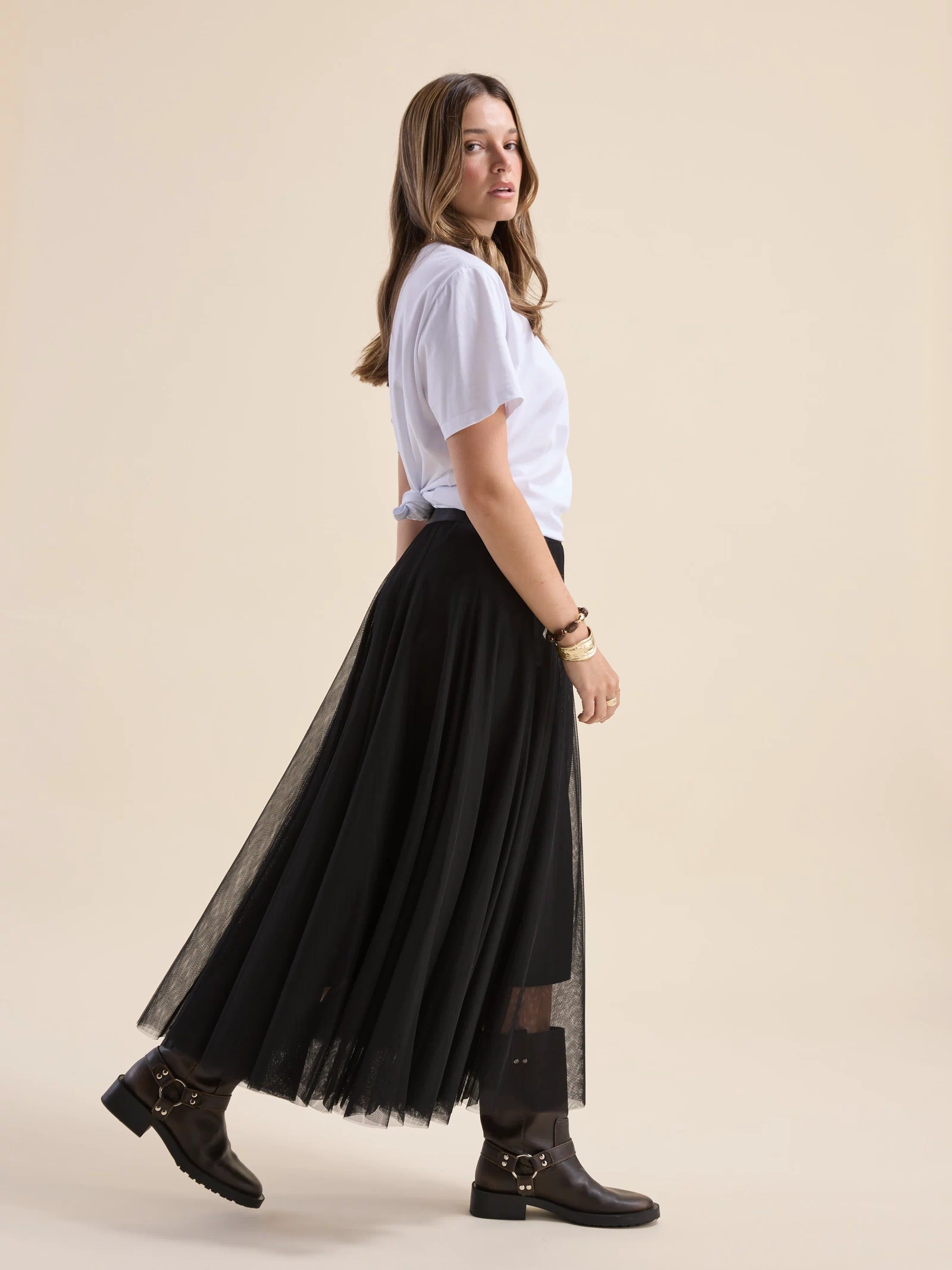 Woman in white tee, Emilie Tulle Skirt by Max, and boots walking sideways on beige background.