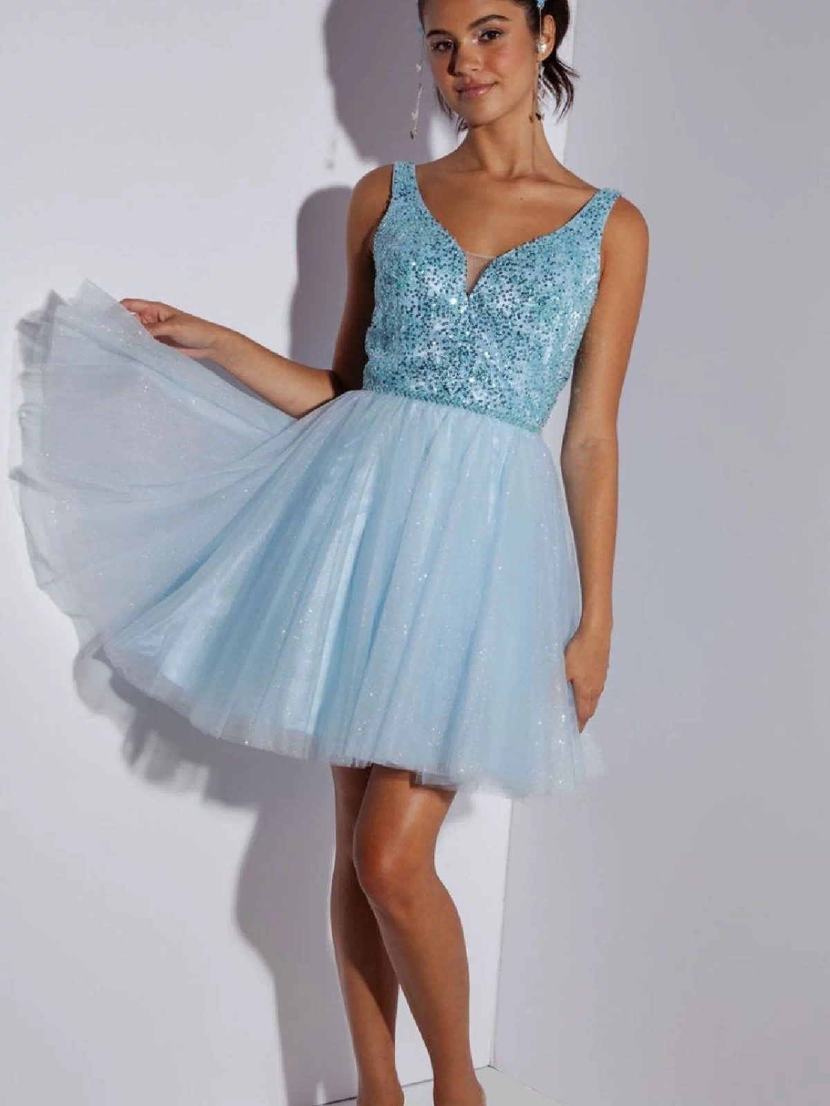 A young woman stands against a white wall, wearing the Eureka 9926 Glitter-Bodice Short A-Line Hoco Dress featuring a sleeveless sequin bodice, light blue tulle skirt, and clear high-heeled shoes.