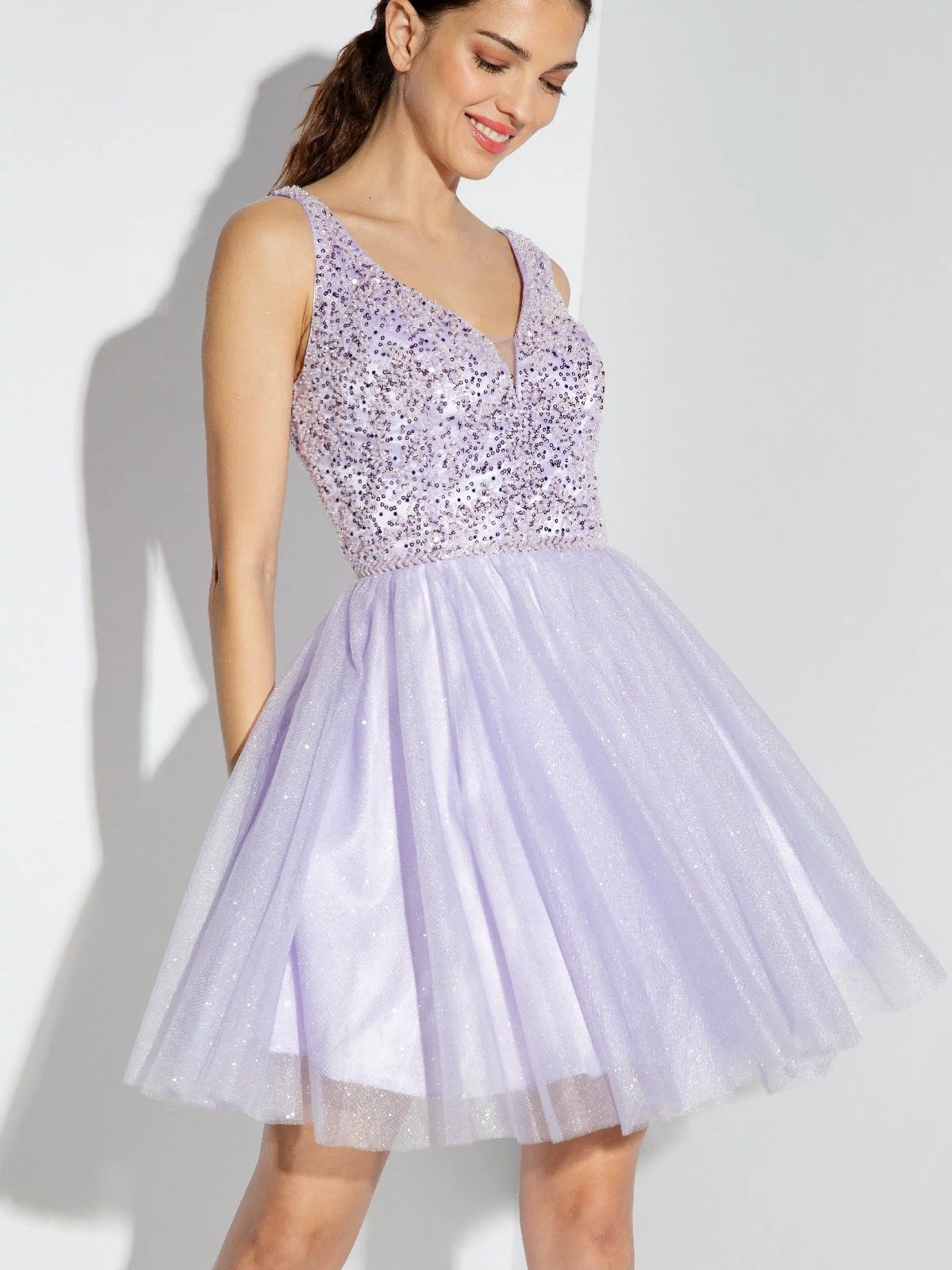 A woman in the Eureka 9926 Glitter-Bodice Short A-Line Hoco Dress, featuring a sleeveless sequin bodice and flared light purple tulle skirt, stands against a white background.