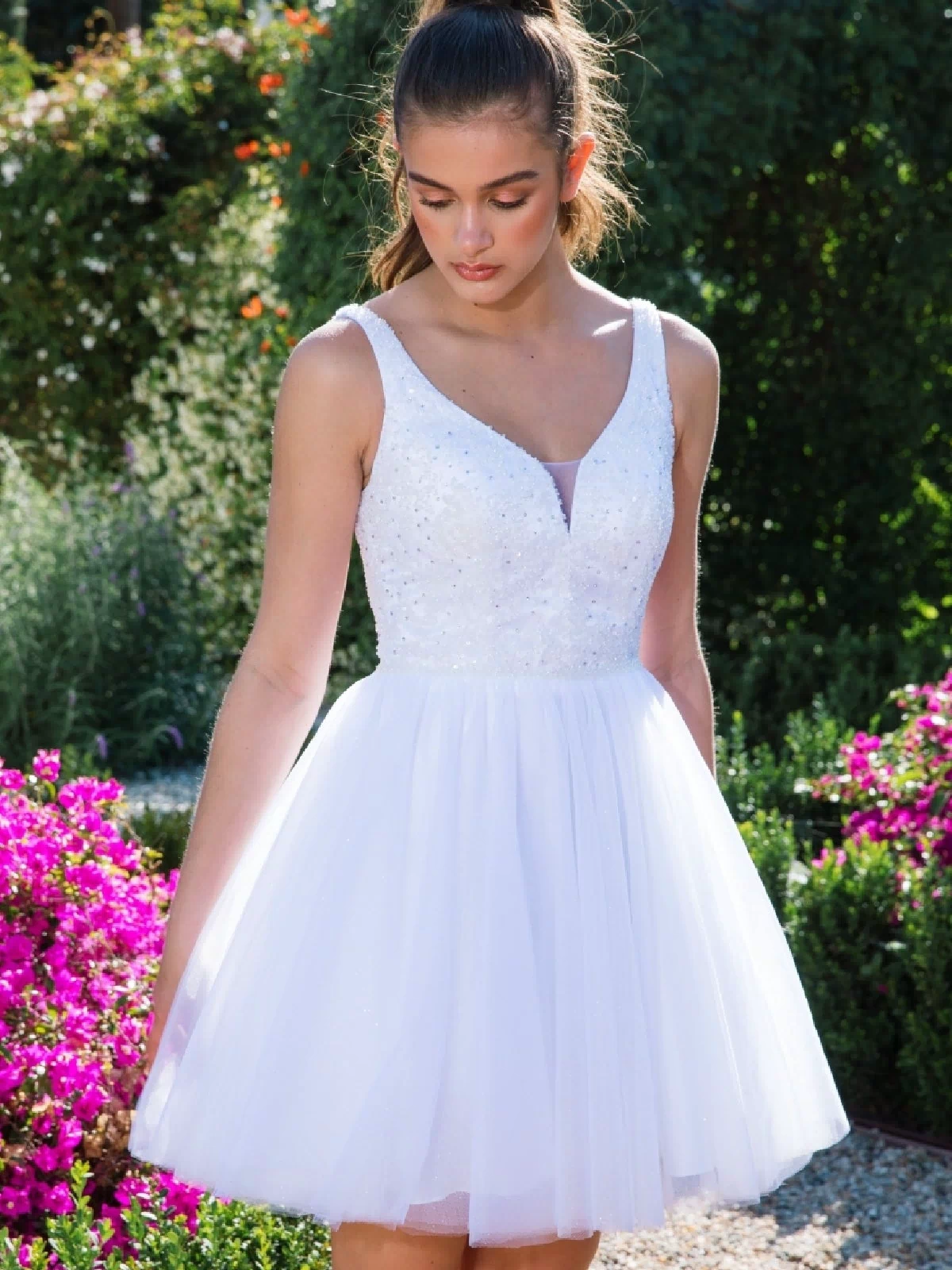 A young woman wears the Glitter-Bodice Short A-Line Hoco Dress: Eureka 9926, featuring a sleeveless white bodice and tulle skirt, as she stands in a blooming garden surrounded by lush greenery.