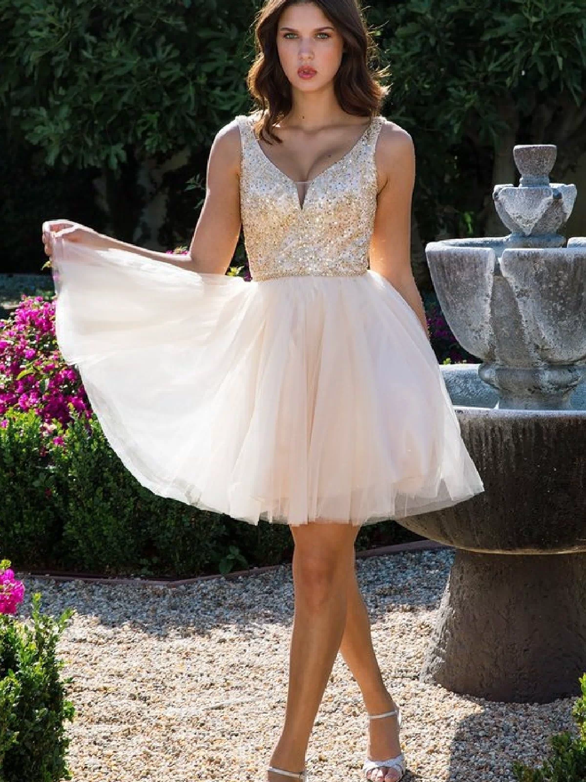 A woman in the Glitter-Bodice Short A-Line Hoco Dress (Eureka 9926), featuring a sleeveless, sequin bodice, stands outdoors near a stone fountain surrounded by lush greenery.