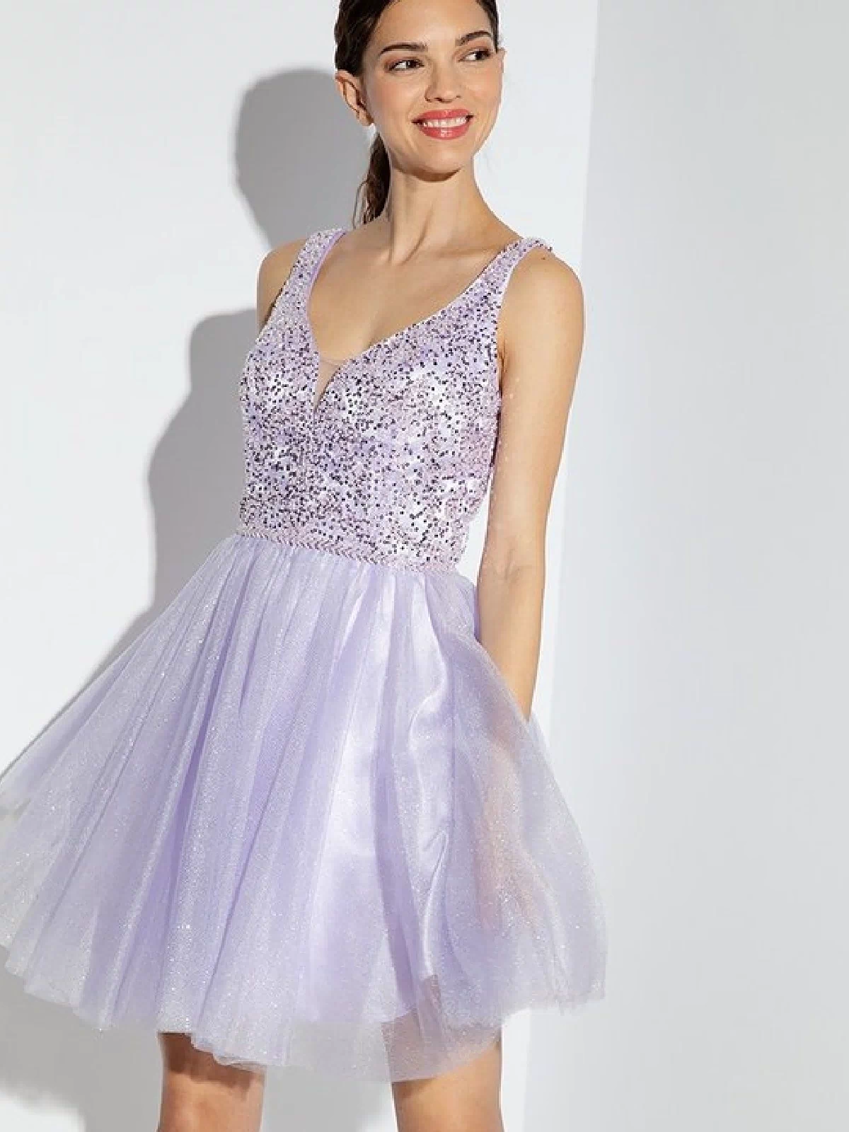 A woman smiles and looks to the side, modeling the Eureka 9926 Glitter-Bodice Short A-Line Hoco Dress—a sleeveless lilac cocktail dress with a sequin bodice and tulle skirt—against a white background.
