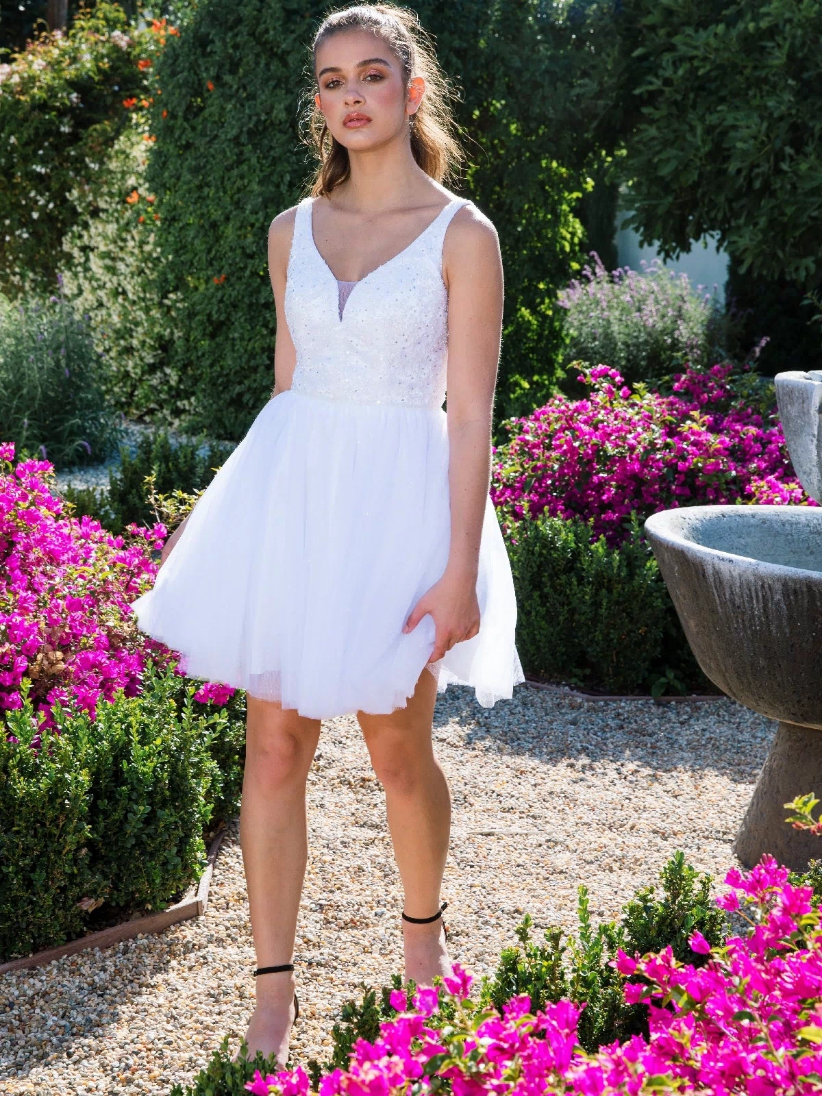 A young woman in the Glitter-Bodice Short A-Line Hoco Dress: Eureka 9926 stands on a garden gravel path surrounded by blooming pink flowers and lush greenery.