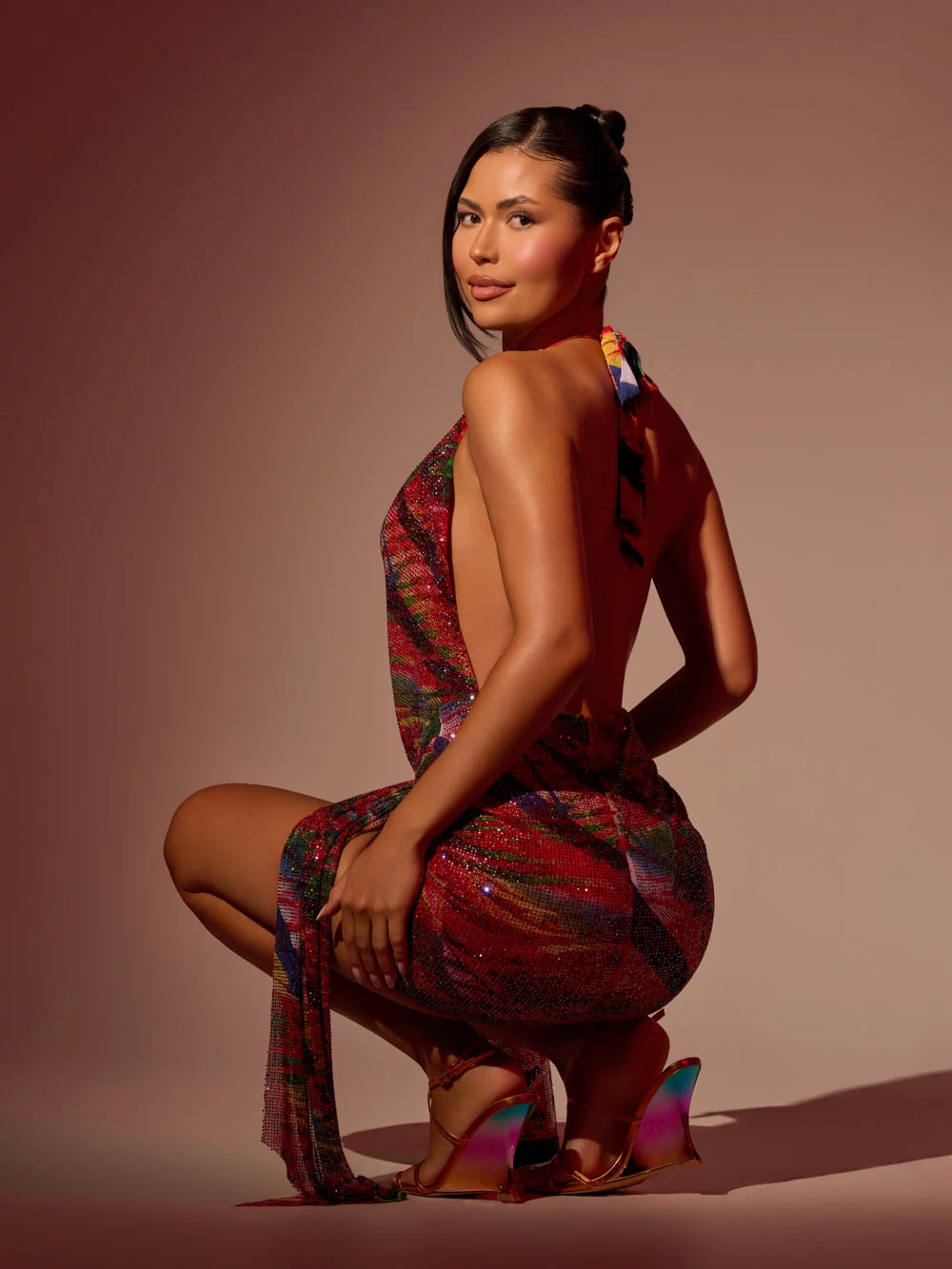 A woman with dark hair in a sleek updo squats against a neutral gradient background, wearing the FEVER DREAM RHINESTONE MINI DRESS and shiny high-heeled sandals.