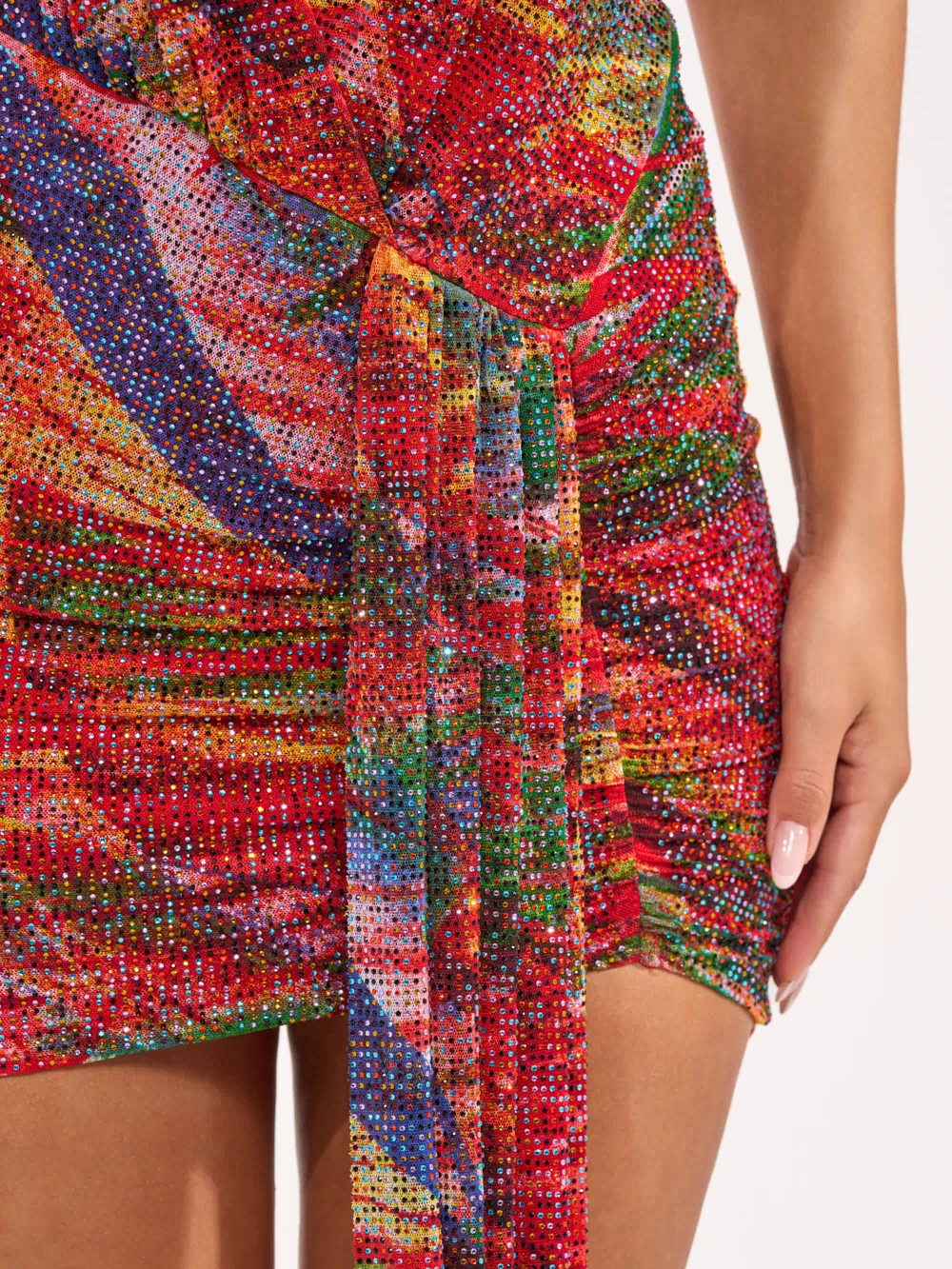 A close-up shows someone in the FEVER DREAM RHINESTONE MINI DRESS—a sparkly red halter mini with ruched side and tie detail. Their hand highlights the vibrant textured fabric, making this dress perfect for any festive occasion.