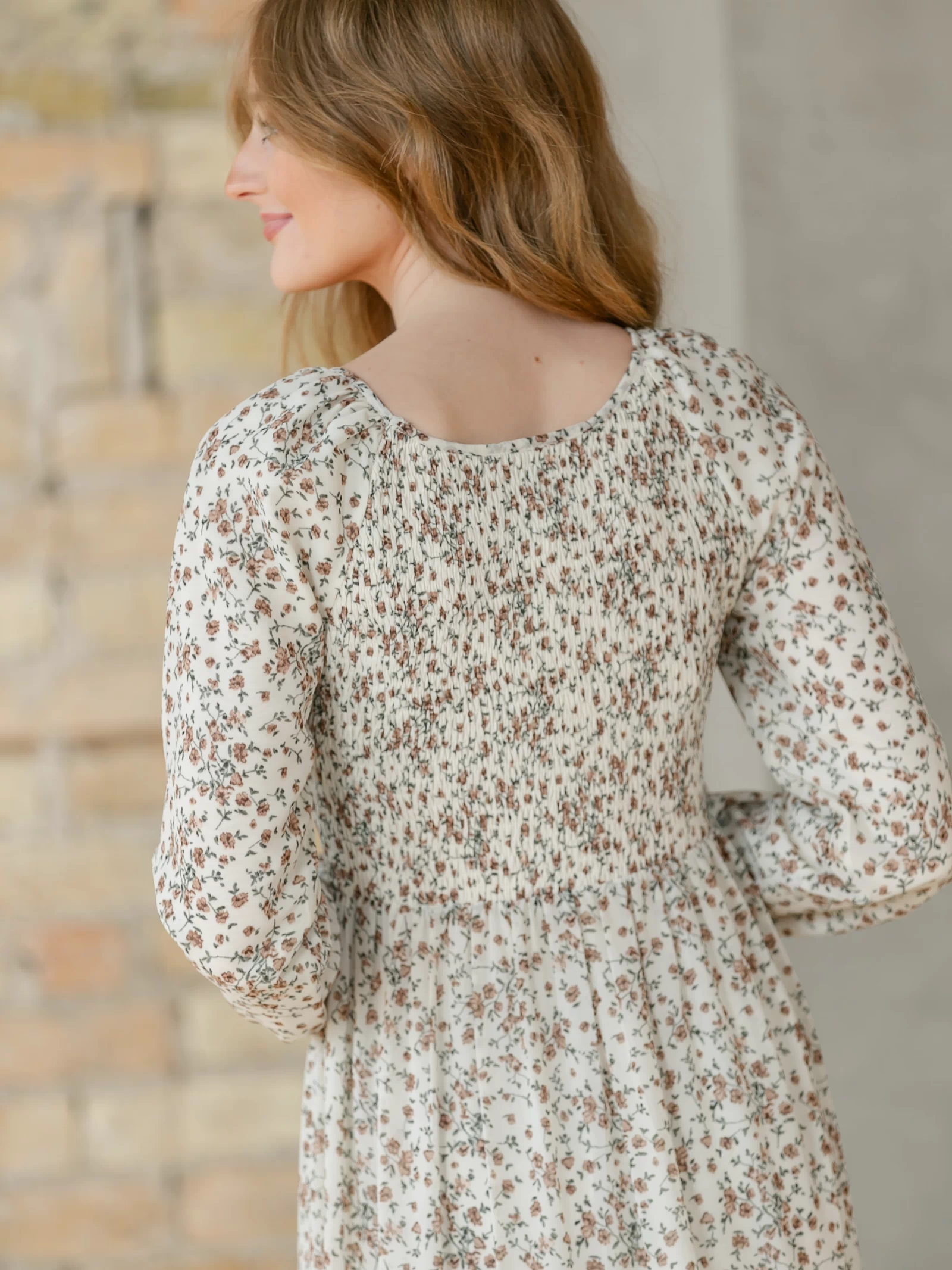 Back view of a woman wearing a floral dress standing against a brick wall.
