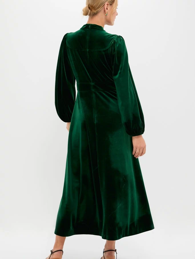 Woman in classic Green Velvet Serena Dress, featuring long balloon sleeves and fitted waist. Effortless elegance with high-end details, styled with black-heeled sandals. Perfect for fun, celebratory events.