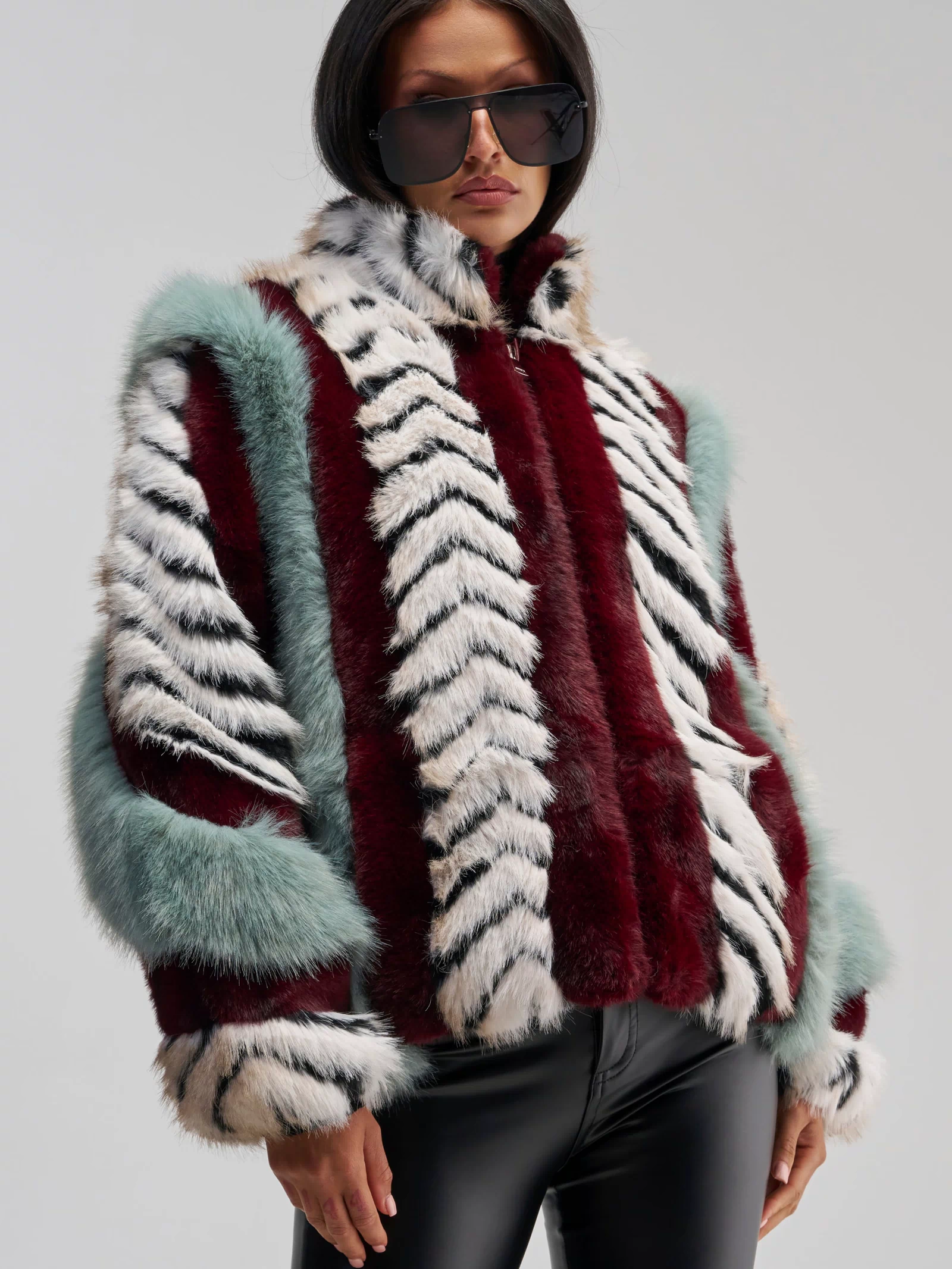 A person wearing large black sunglasses and the HAVE IT ALL PANELED FUR COAT—a multicolored faux fur jacket featuring red, white, black, and teal stripes in an oversized silhouette—paired with shiny black pants stands against a plain, light gray background.