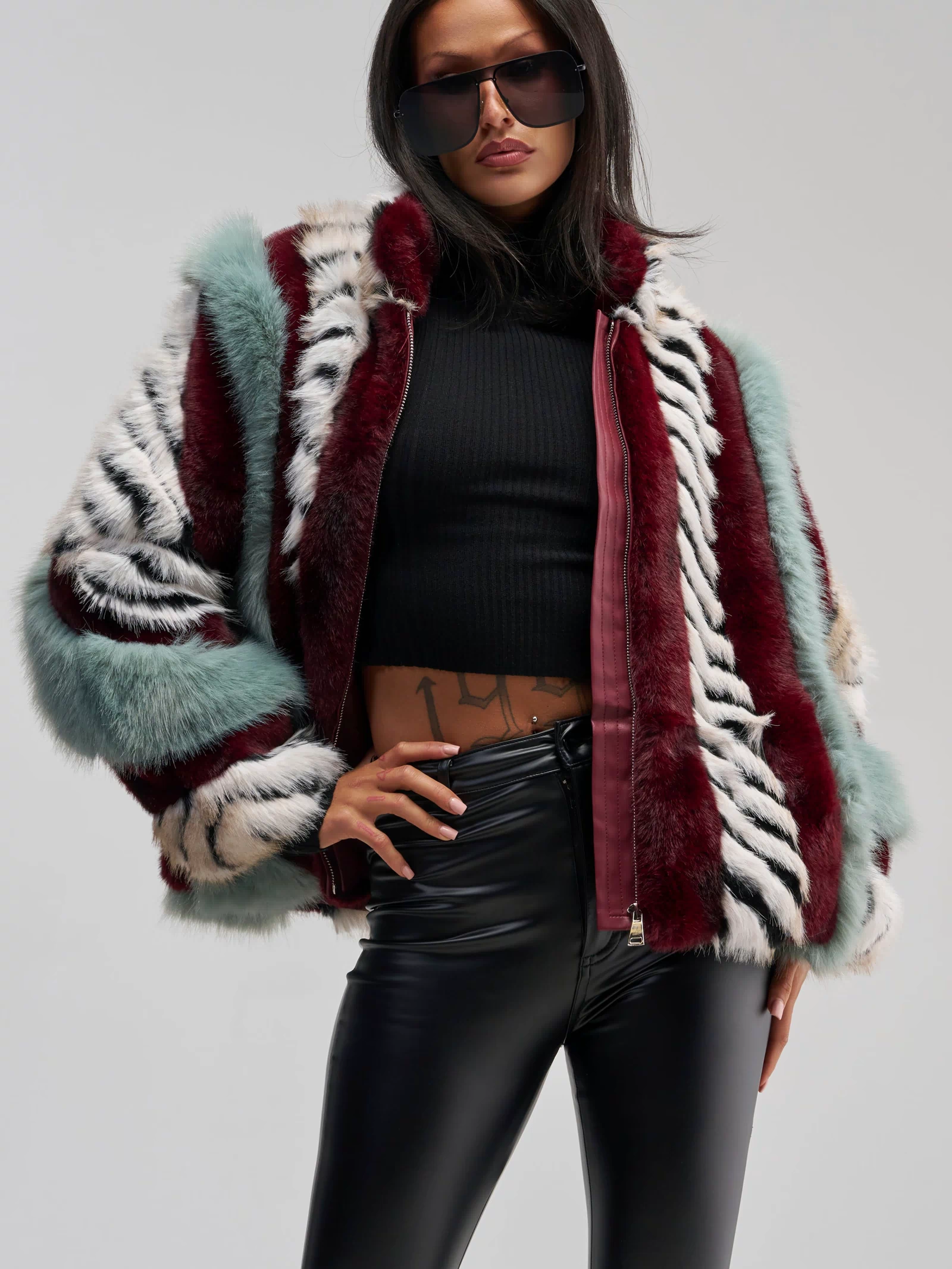 A woman in large sunglasses wears the HAVE IT ALL PANELED FUR COAT—a multicolored faux fur jacket with zebra stripes—styled with a black crop top and black leather pants, standing confidently with one hand on her hip.