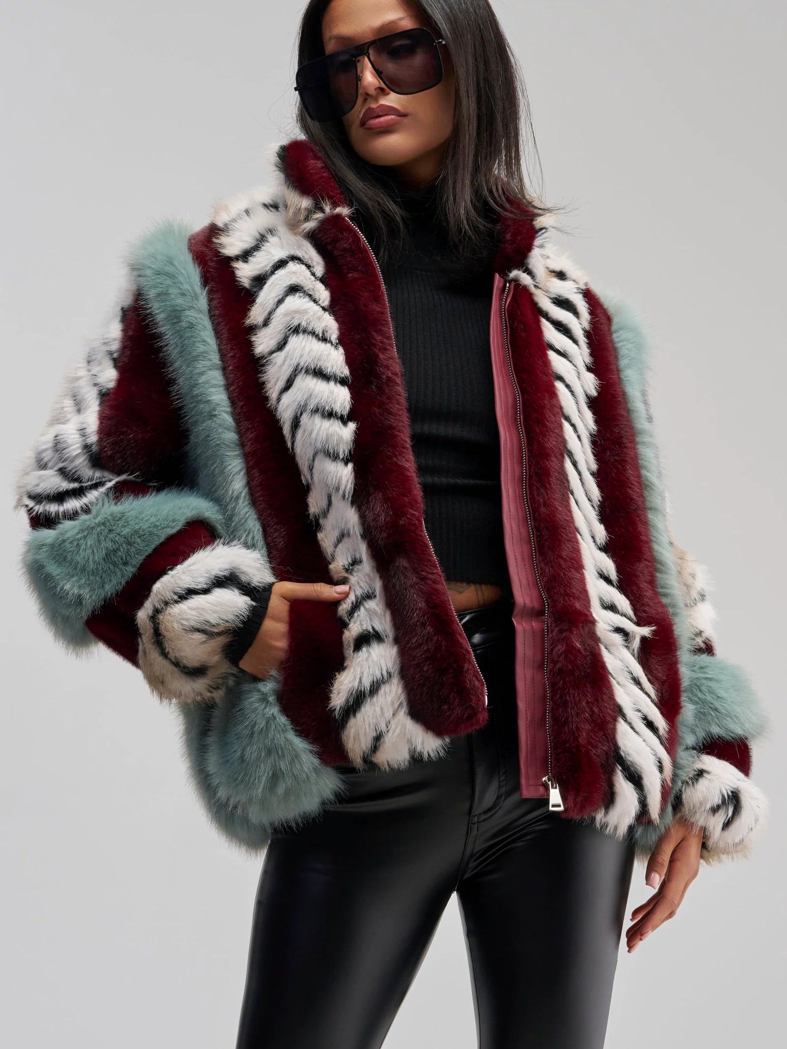 A woman poses against a neutral background wearing the HAVE IT ALL PANELED FUR COAT—a colorful faux fur jacket with an oversized silhouette and red, white, and teal paneled patterns—styled with a black turtleneck, black leather pants, large sunglasses, and one hand in her pocket.