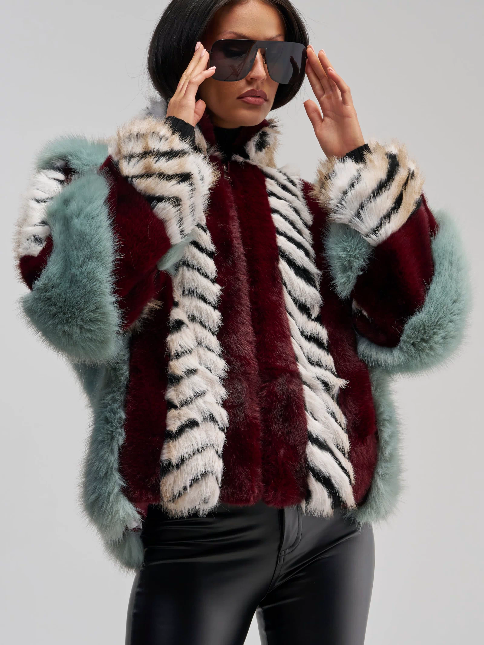 A woman poses confidently against a plain background wearing the HAVE IT ALL PANELED FUR COAT—an oversized faux fur jacket featuring multicolored panels with striped accents and teal sleeves—styled with black leather pants and large sunglasses.