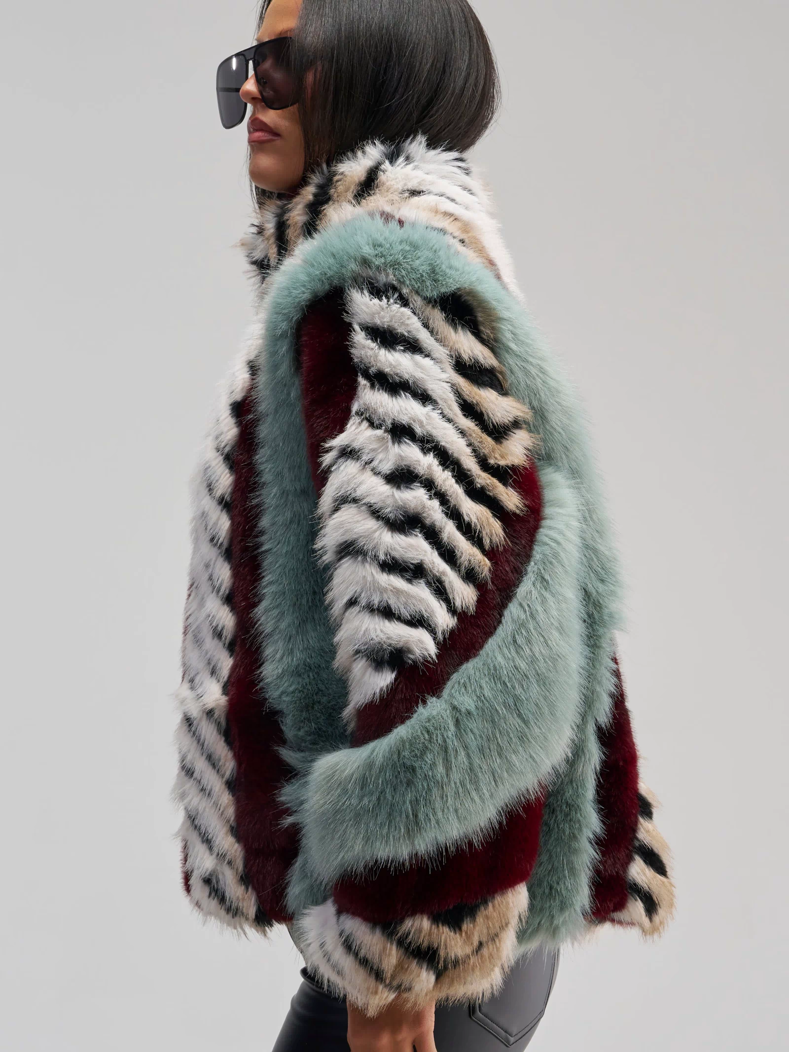 Wearing the HAVE IT ALL PANELED FUR COAT—an oversized faux fur jacket featuring bold teal, burgundy, black, and white stripes—a person in large black sunglasses stands sideways against a plain background.