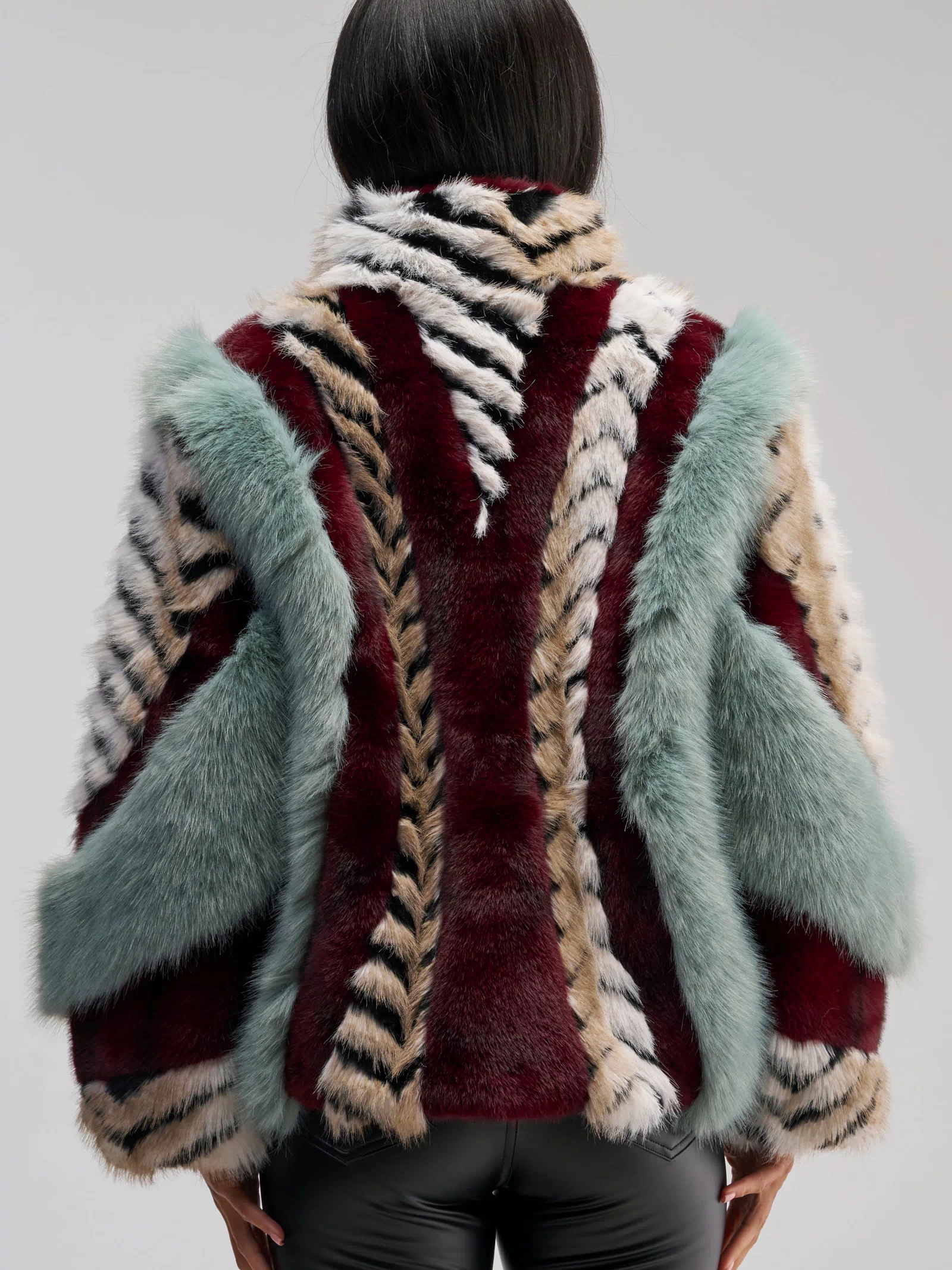 A person with straight dark hair is photographed from behind against a plain background, wearing the HAVE IT ALL PANELED FUR COAT—an oversized faux fur jacket featuring multicolored panels in burgundy, cream, black, and teal—paired with black leather pants.
