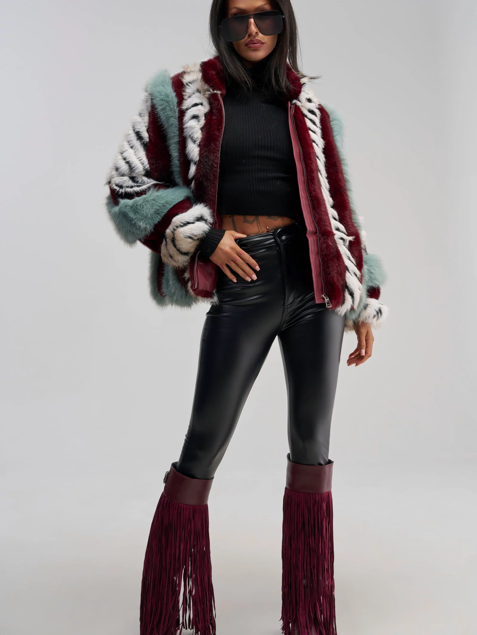 A woman stands confidently against a plain background, wearing the HAVE IT ALL PANELED FUR COAT—a multicolored faux fur jacket in an oversized silhouette—styled with a black crop top, black leather pants, and maroon fringe boots.