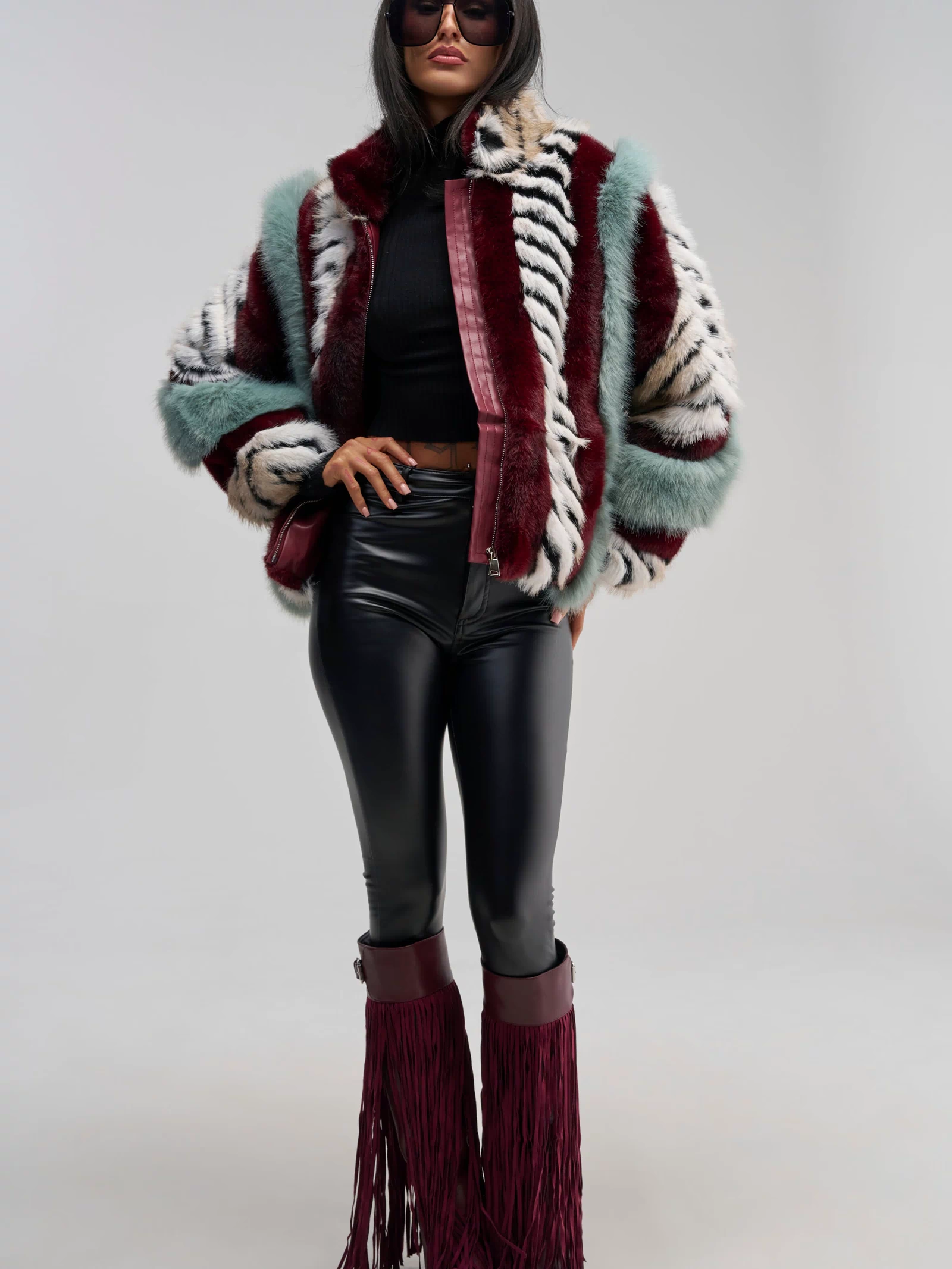 A person wearing the HAVE IT ALL PANELED FUR COAT—featuring a multicolored paneled faux fur and an oversized silhouette—paired with a black crop top, black leather pants, burgundy fringe boots, and sunglasses stands against a plain gray background.
