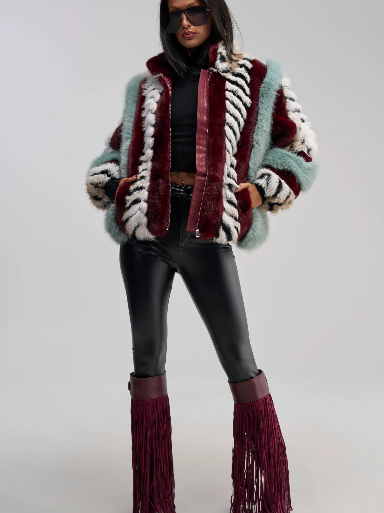 A woman in sunglasses wears the HAVE IT ALL PANELED FUR COAT—a multicolored, paneled faux fur jacket with an oversized silhouette—styled with a black crop top, black leather pants, and maroon fringed boots as she stands confidently against a plain background.