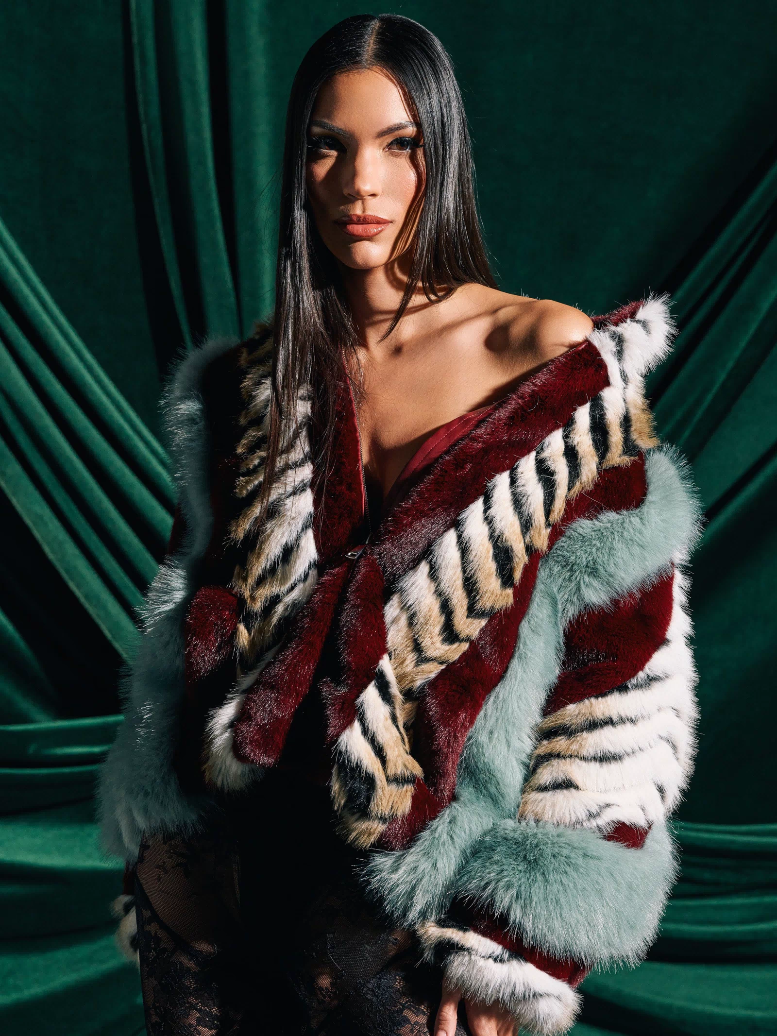A woman with long straight hair poses before dark green drapes, wearing the HAVE IT ALL PANELED FUR COAT featuring bold red, white, black, and pale blue stripes over a black lace outfit.