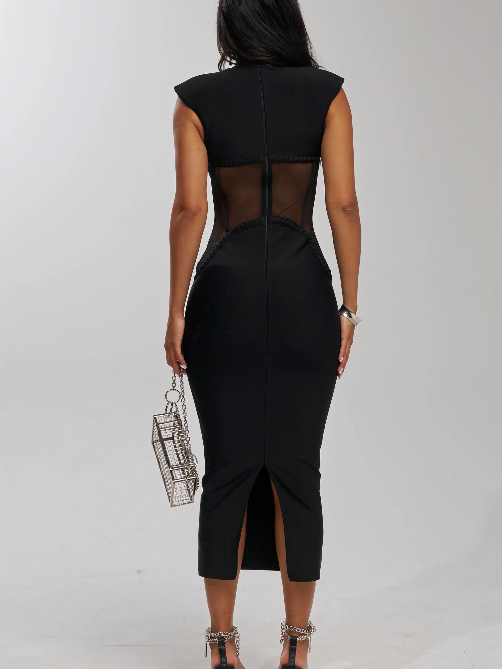 A woman faces away, wearing the ICONIC INSTINCT BANDAGE CORSET MIDI DRESS with sheer back panels, black chain-detailed heels, and holding a small silver boxy purse with a chain strap. The background is plain and light-colored.