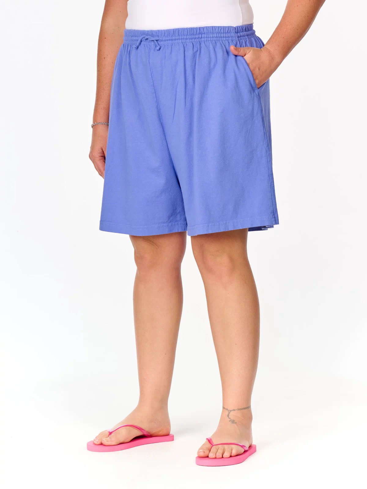 A person wearing a white tank top, the Fresh Produce Clothes Jersey Shorts | Plus Size in light blue with deep side pockets, and pink flip-flops, standing against a white background.
